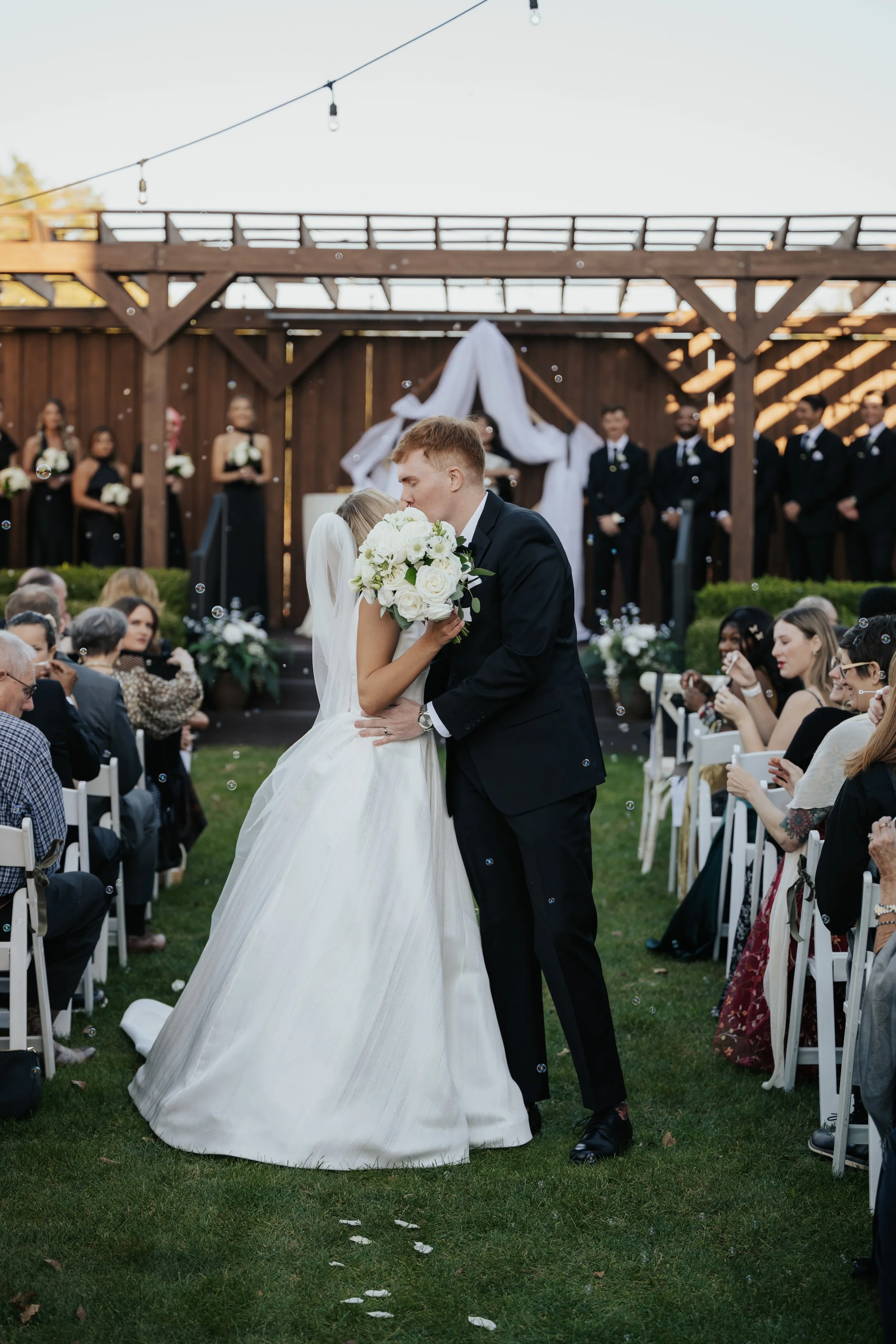A bride and groom share a kiss at their outdoor wedding ceremony. The bride wears a white gown and veil; the groom is in a black tuxedo. Guests sit on either side, and bridesmaids and groomsmen stand in the background. Garden decor and a wooden structure with draped fabric are visible.