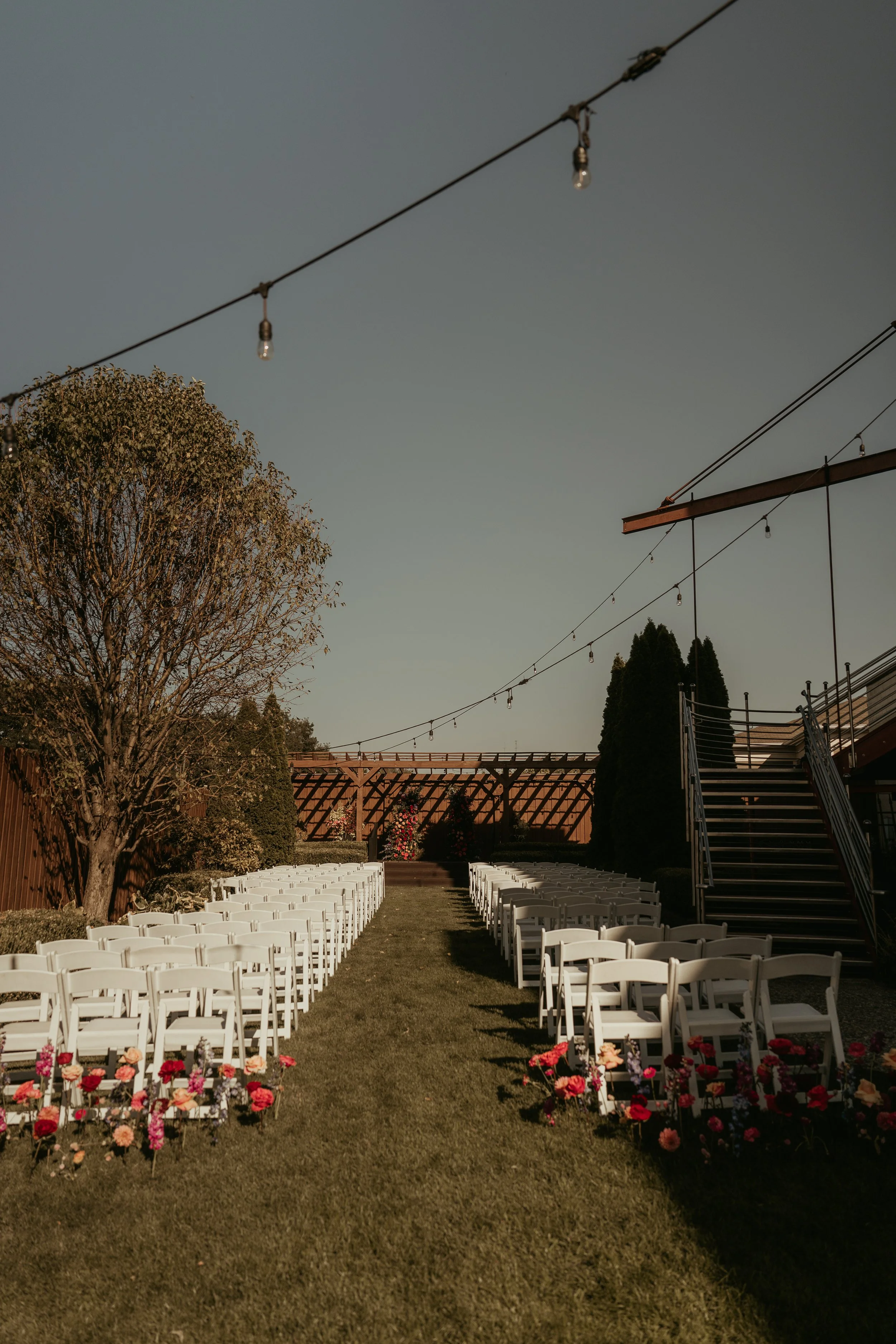 Outdoor wedding ceremony setup with white chairs arranged in rows on either side of an aisle, decorated with colorful flowers in the foreground. Tall trees and a wooden pergola or arbor in the background. Overhead string lights hanging across the sce