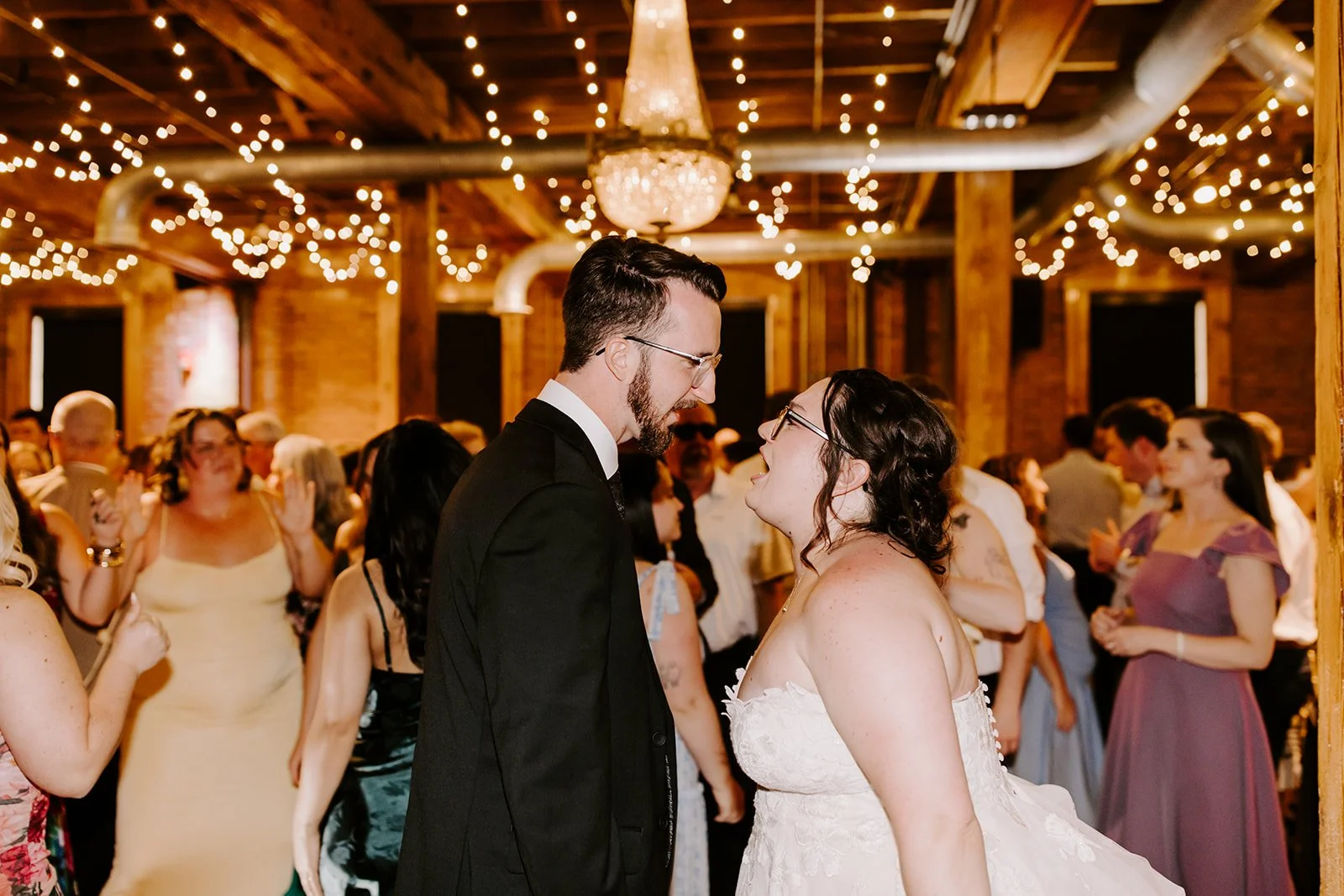 A wedding reception scene with a bride and groom facing each other, inside a decorated venue with warm lighting and string lights overhead.