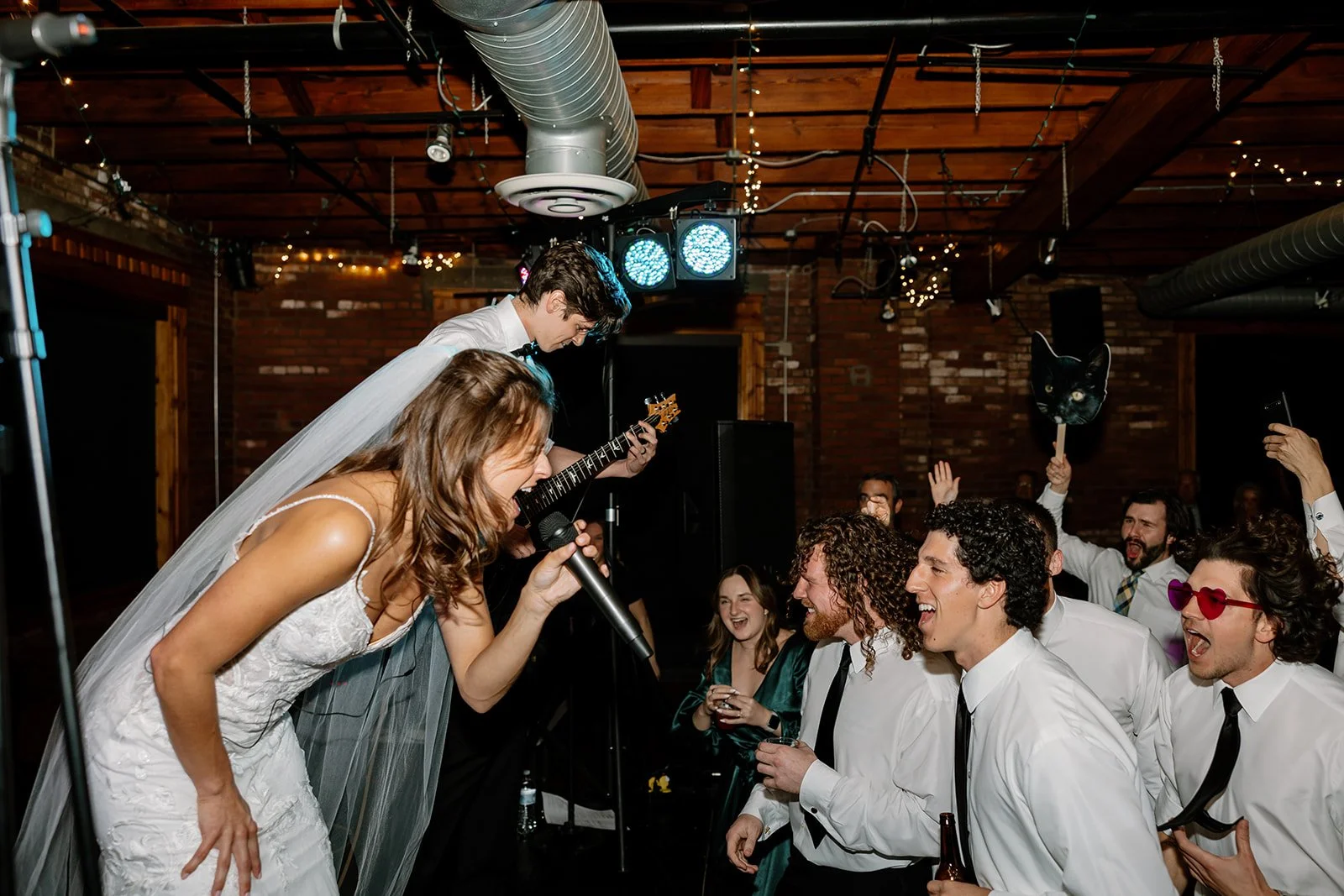 People at a celebration, with a woman singing into a microphone, a man playing guitar, and others dancing and enjoying in a decorated indoor venue with string lights.