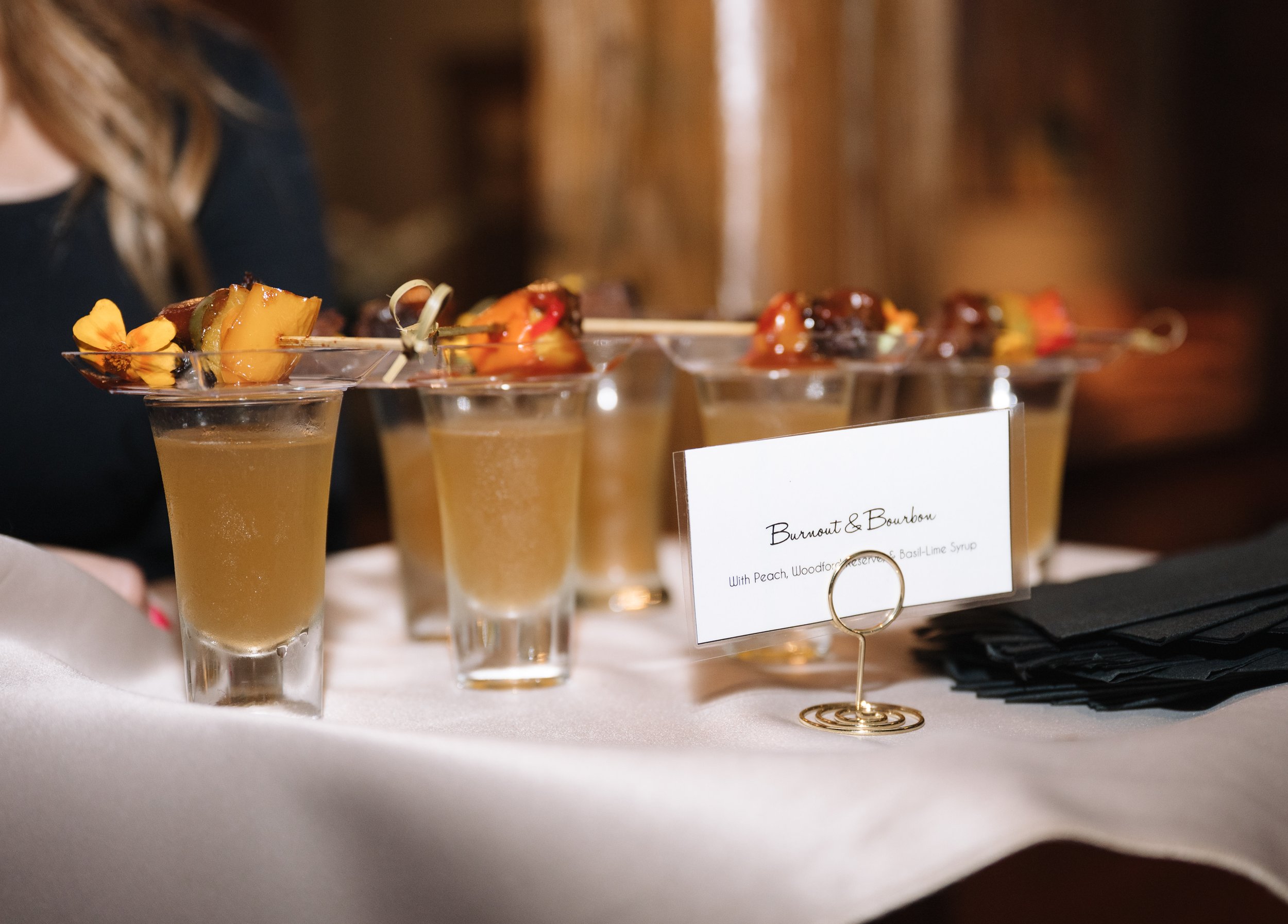 A row of cocktail glasses filled with a yellow-orange beverage garnished with pineapple, cherry, and herbs, on a white table at a tasting event.