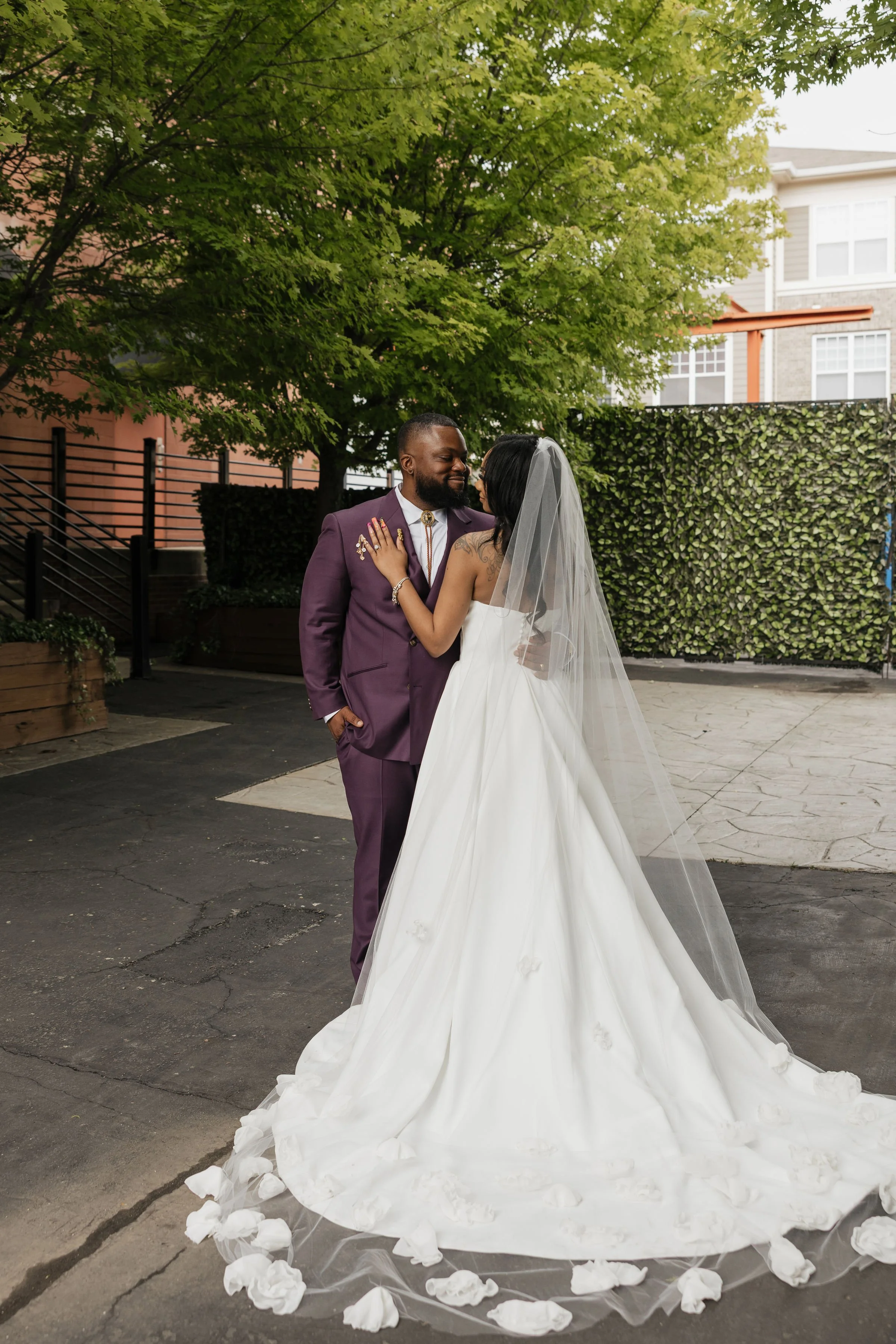 A bride and groom facing each other under a large tree during their wedding, with the bride wearing a white wedding gown and veil, and the groom in a purple suit, in an outdoor urban setting.