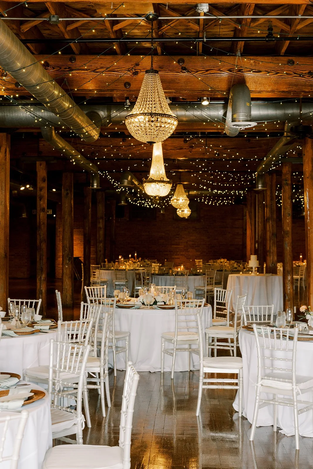 Elegant wedding reception with round tables draped in white tablecloths, white Chiavari chairs, floral centerpieces, and silverware. Crystal chandeliers and string lights adorn the wooden ceiling, creating a warm ambiance in a rustic venue with exposed brick walls.
