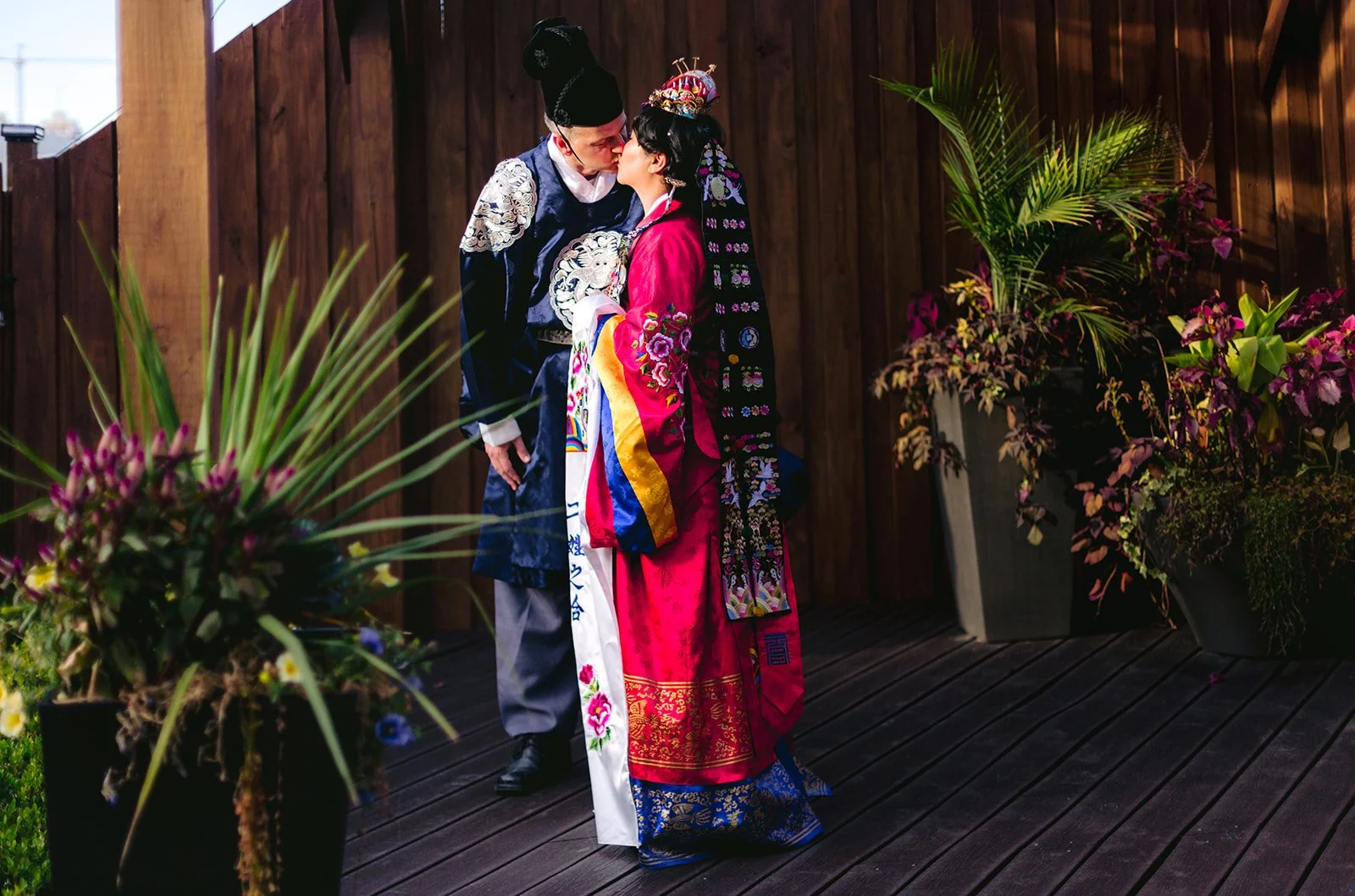 A couple in traditional Korean wedding attire sharing a kiss on a wooden deck with potted plants and a wooden fence in the background.
