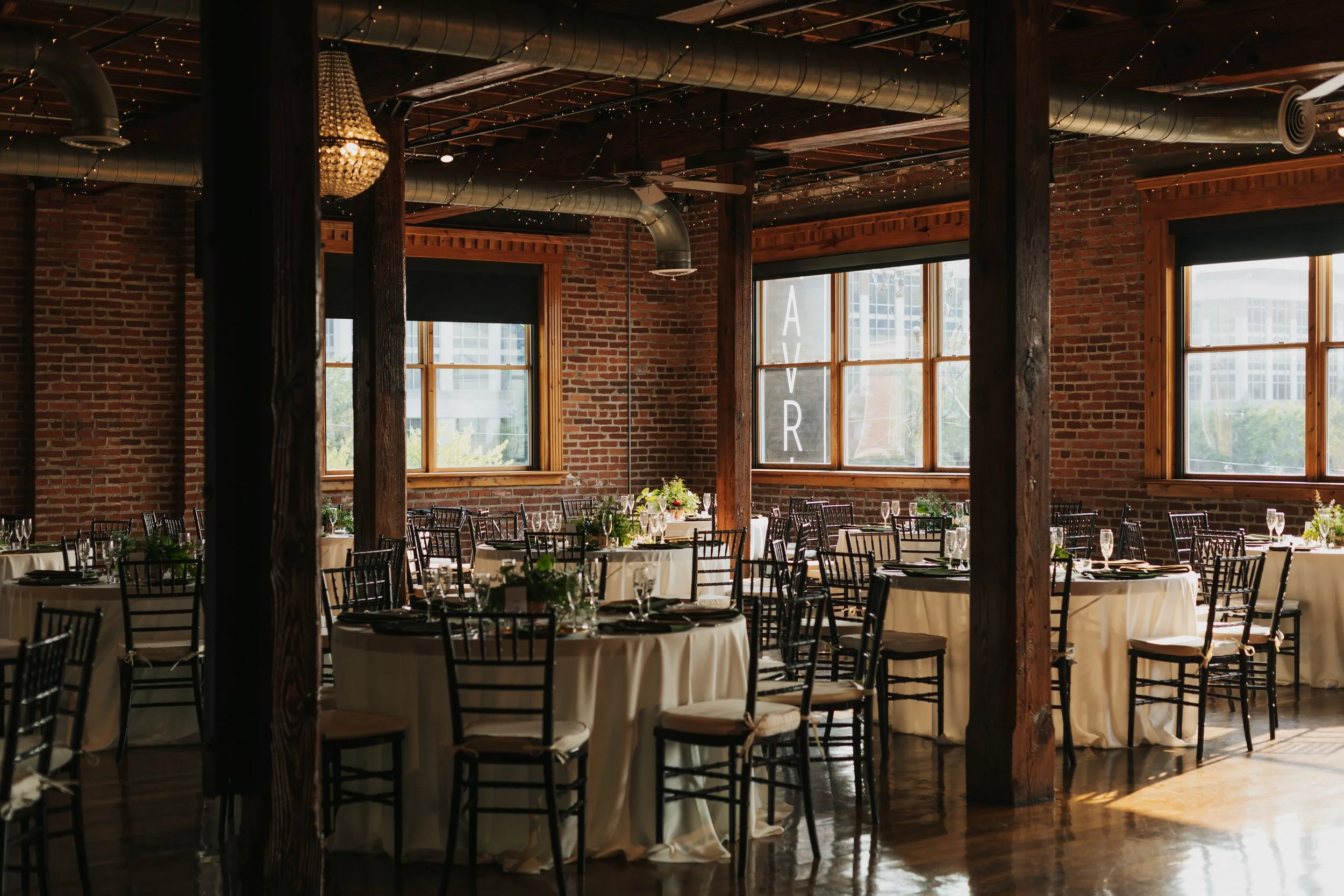 Indoor event space with brick walls, large windows, wooden beams, chandeliers, string lights, and round tables set for a meal with chairs.