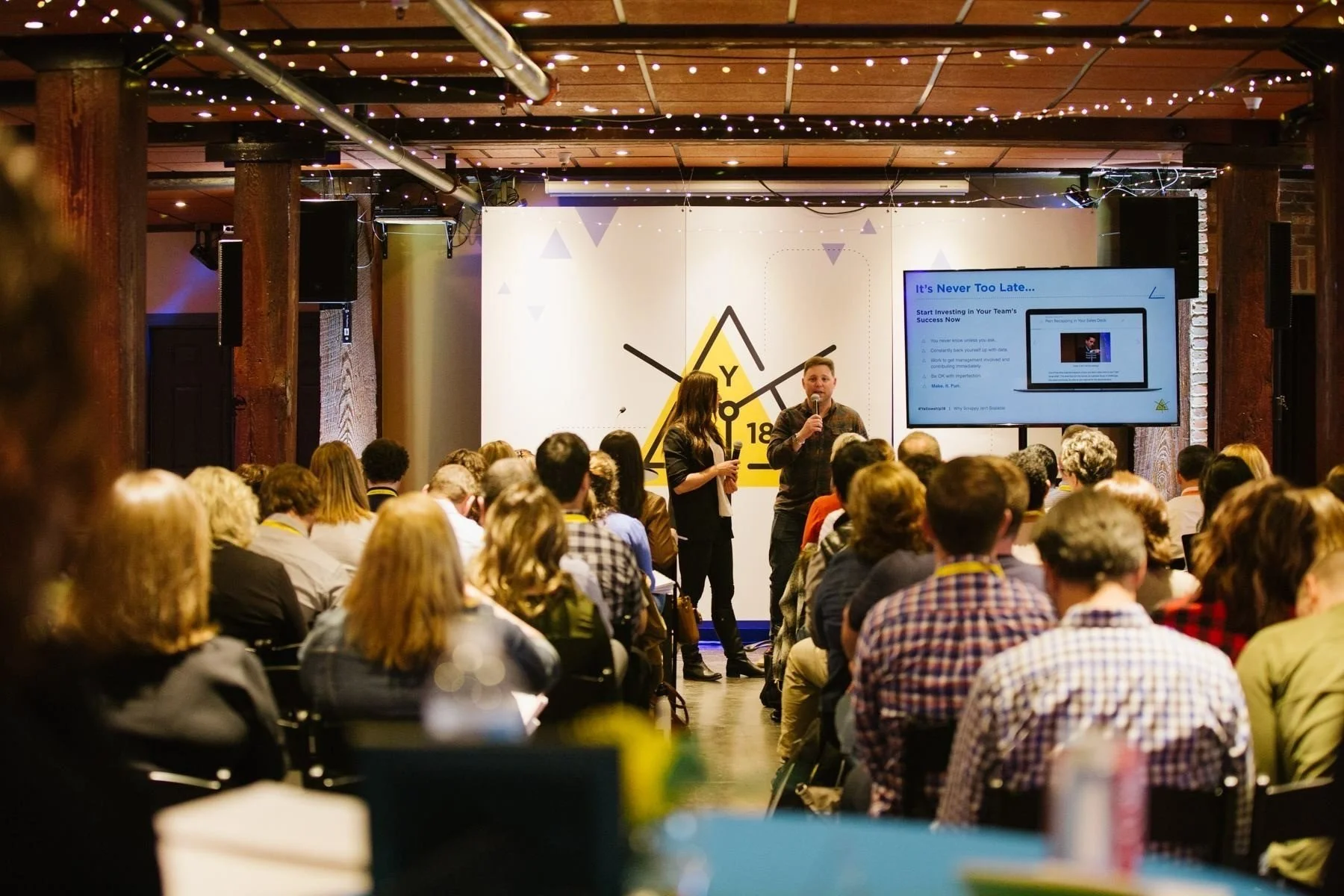 A conference or seminar room filled with people sitting and listening to a presentation. Two speakers are on stage, one man and one woman, with a large screen displaying a slide titled 'It's Never Too Late...' The room has a wooden ceiling with strin