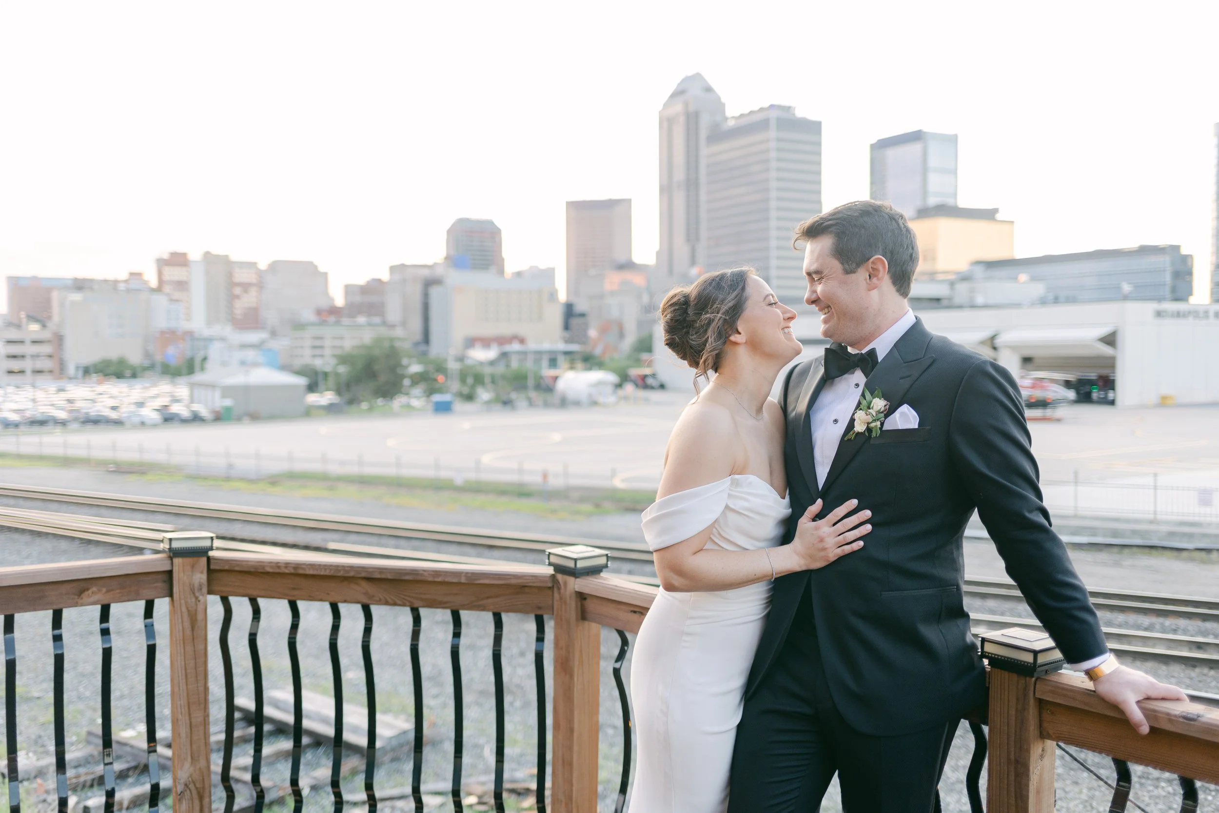A newlywed couple smiling and looking at each other, standing on a wooden deck with a city skyline in the background.