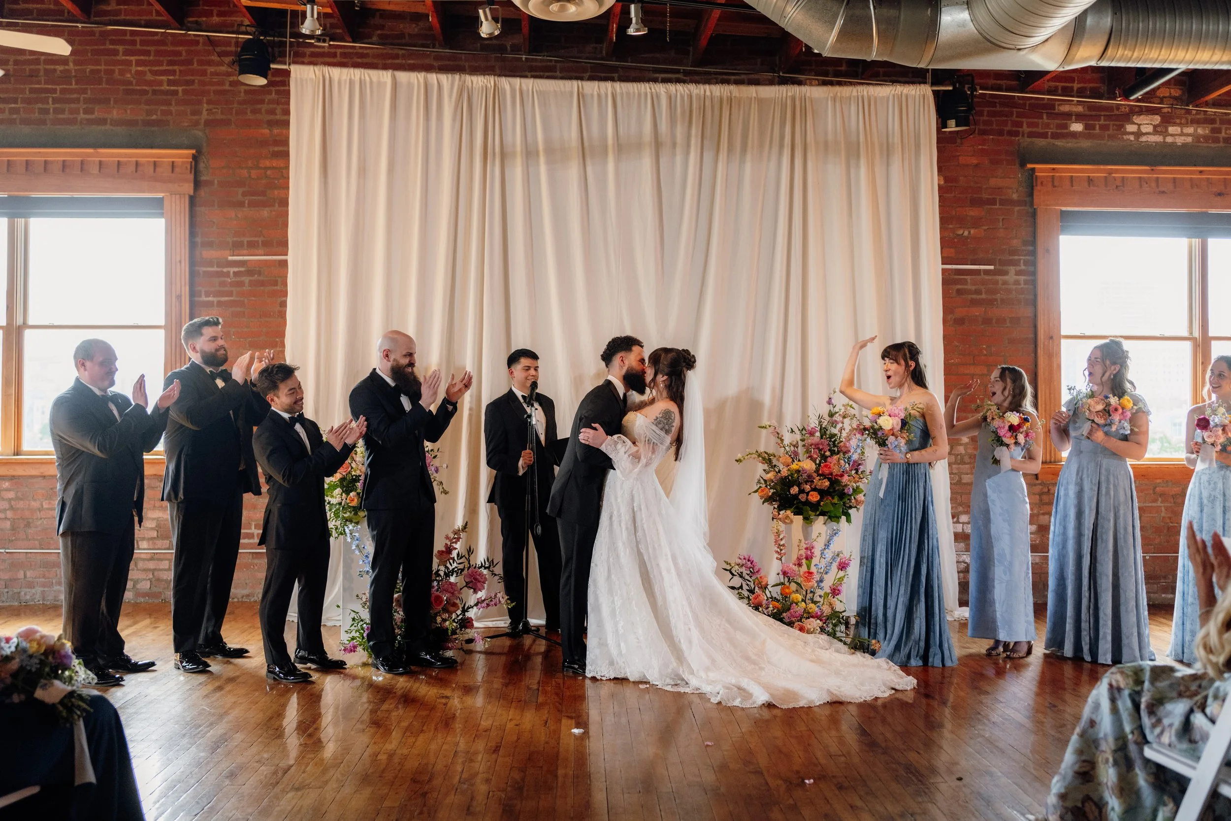 A wedding ceremony with the bride and groom kissing at the altar, surrounded by wedding party members including bridesmaids and groomsmen, with floral arrangements and curtains in the background.