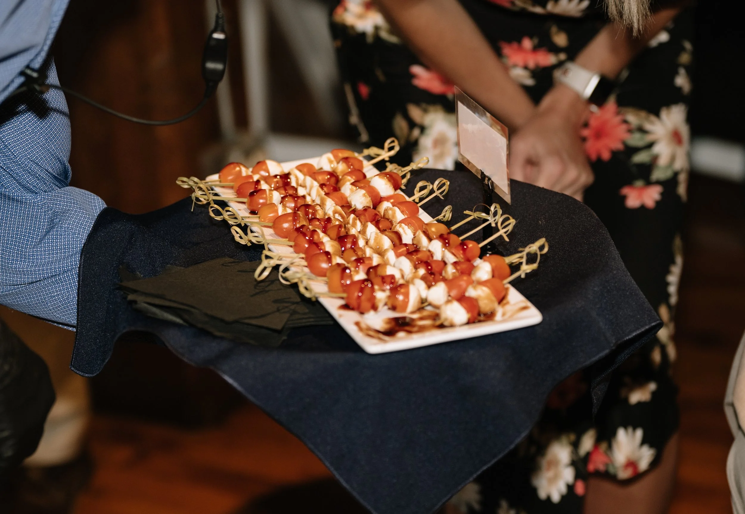 Person holding a black tray with two slices of skewered appetizers topped with ketchup and possibly mayonnaise. Someone wearing floral dress is partially visible in the background.