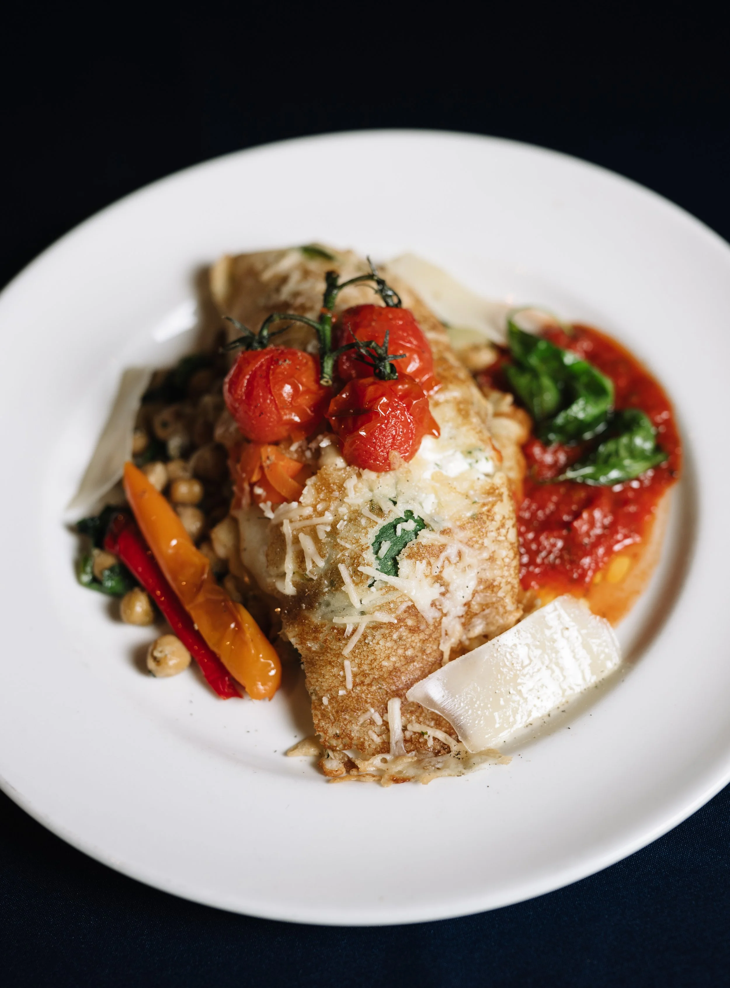 Baked white fish fillet topped with cherry tomatoes on a stem, garnished with grated cheese, served with roasted bell peppers, chickpeas, and greens on a white plate.