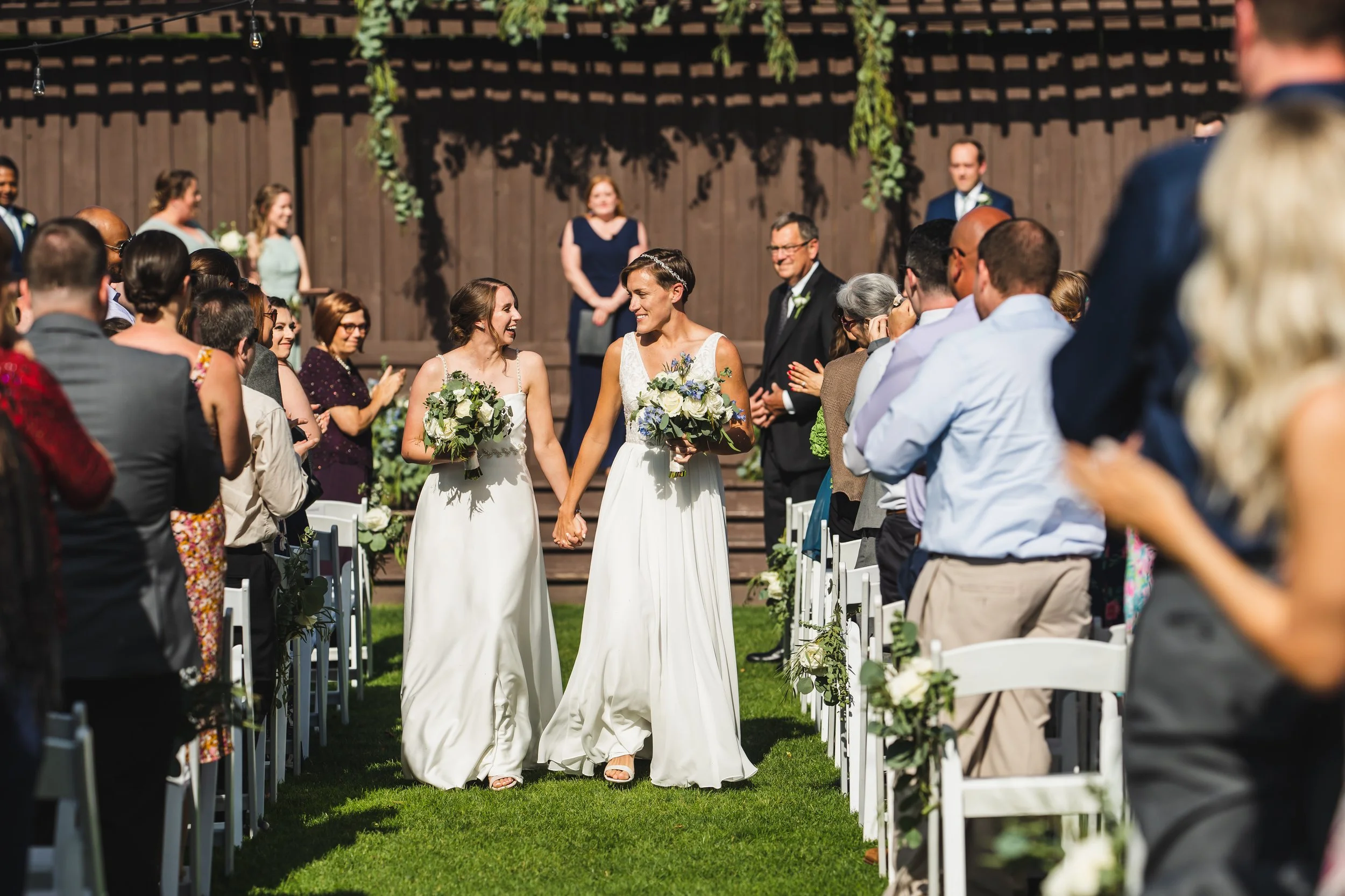 Two brides holding hands and carrying bouquets walking down the aisle at an outdoor wedding ceremony, surrounded by seated guests, with a wooden fence and greenery in the background.