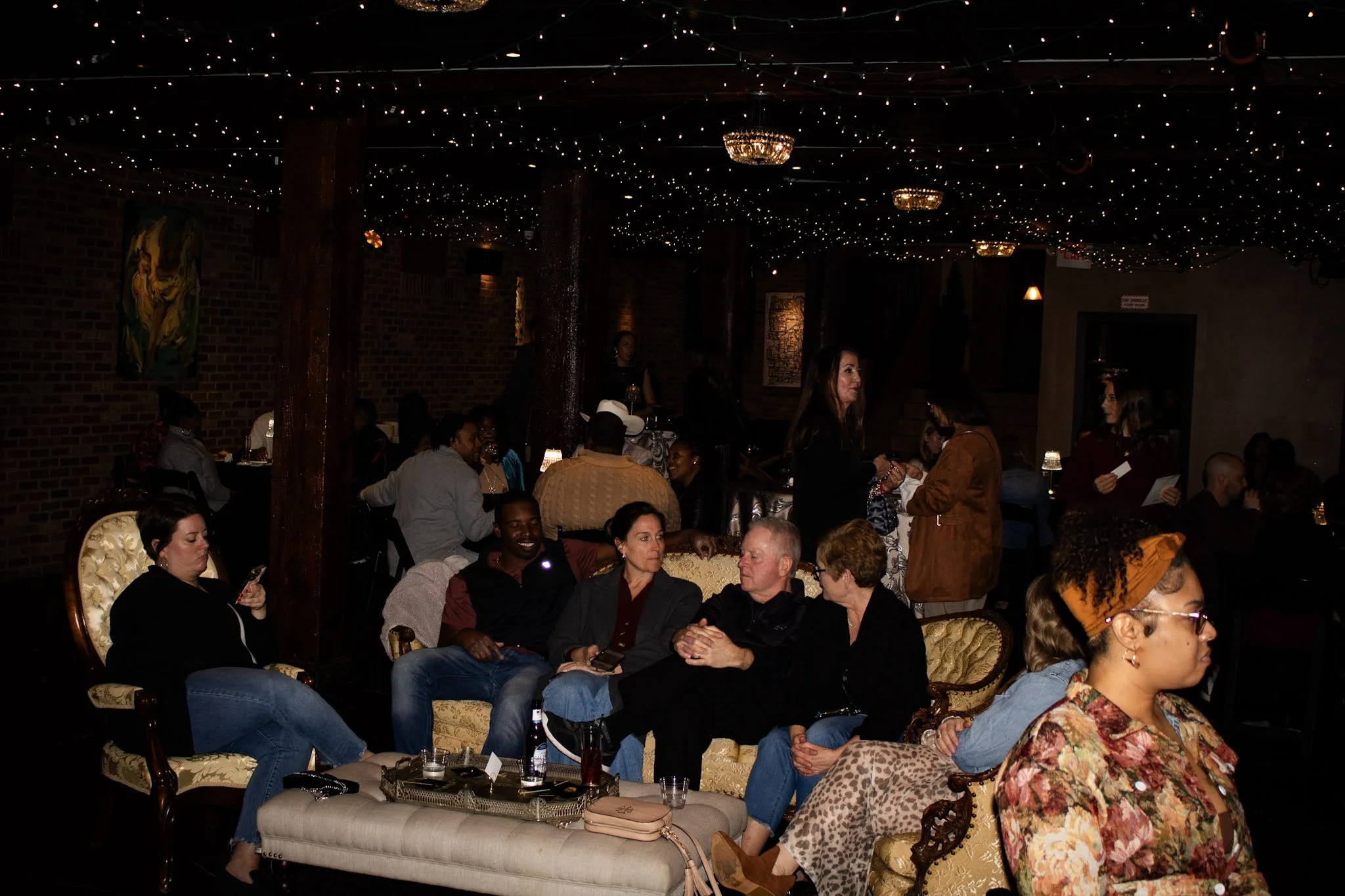People seated at tables in a dimly lit venue with string lights and chandeliers hanging from the ceiling, engaged in conversation and enjoying an event or gathering.