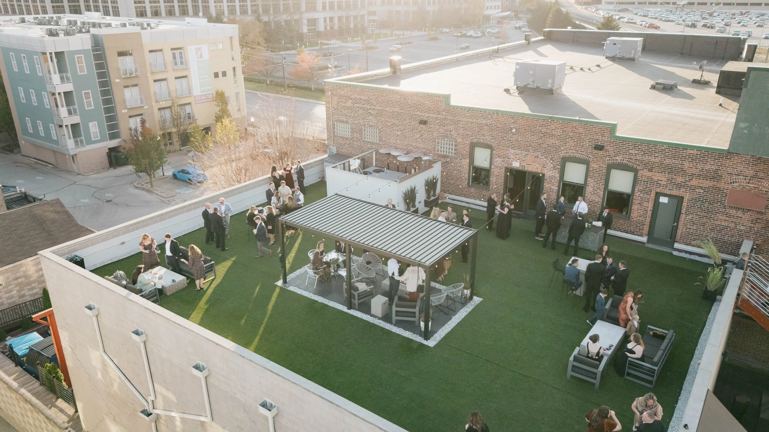 People gathering on a rooftop terrace for an outdoor event, with tables, seating, and string lights, under a clear sky in an urban setting.