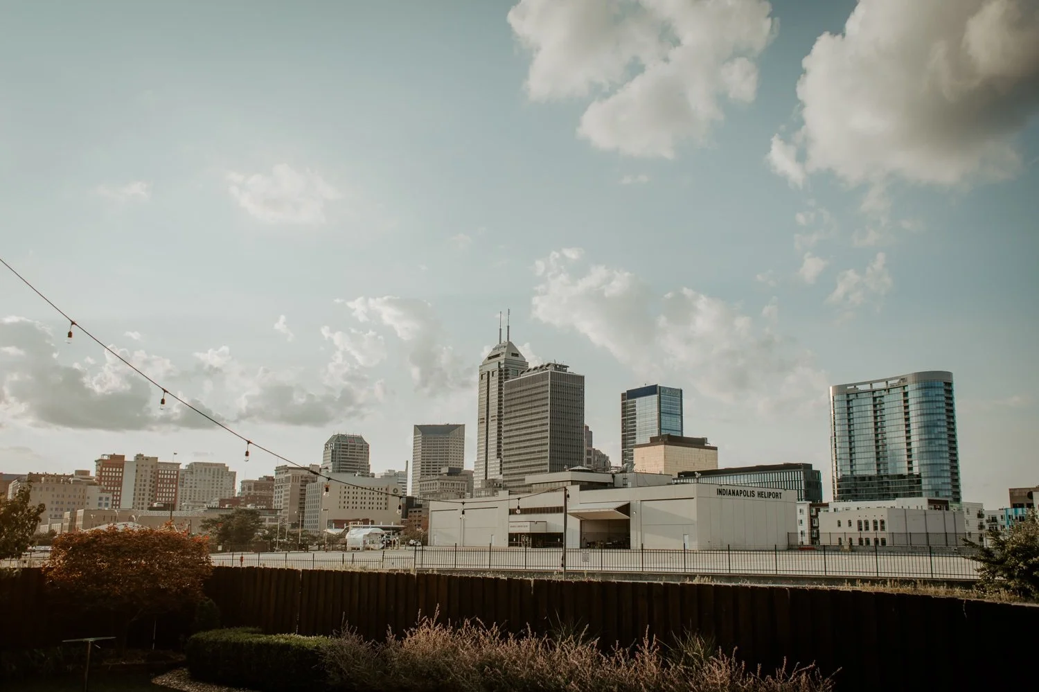 City skyline with tall office buildings and a clear sky, view from a rooftop with string lights and a fenced yard in the foreground.