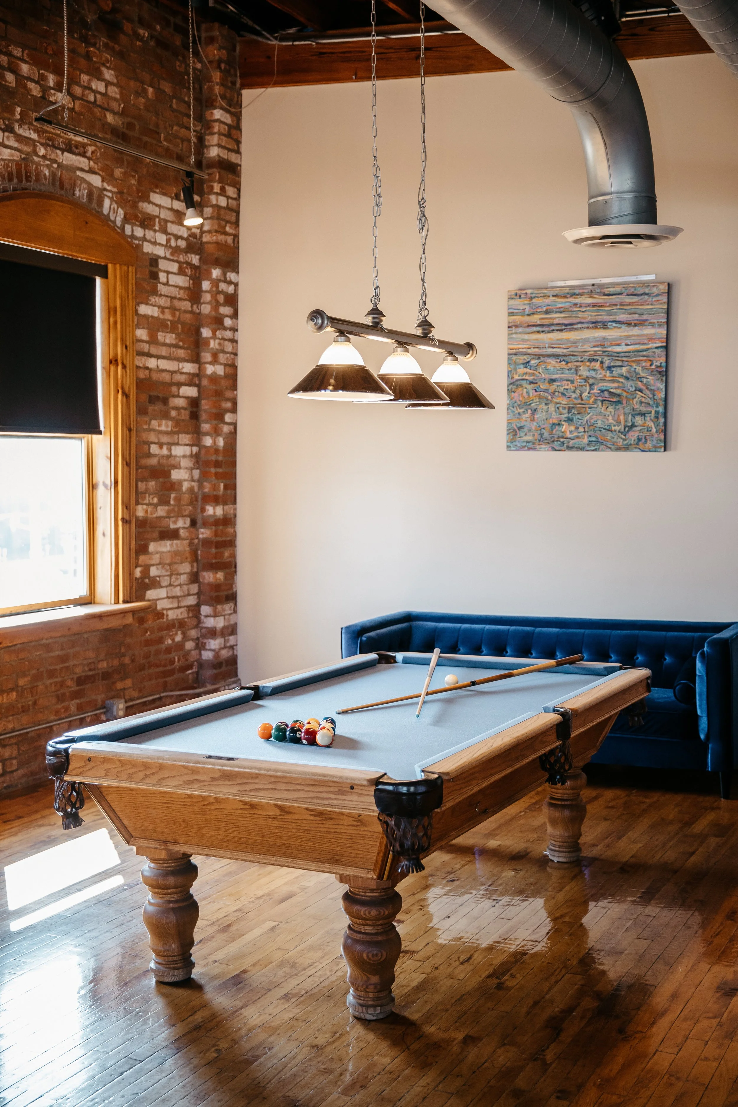 A pool table with billiard balls racked at one end, cue sticks on the table, in a room with exposed brick wall, hardwood floor, a window, a colorful abstract painting, and industrial-style lighting fixtures hanging overhead.