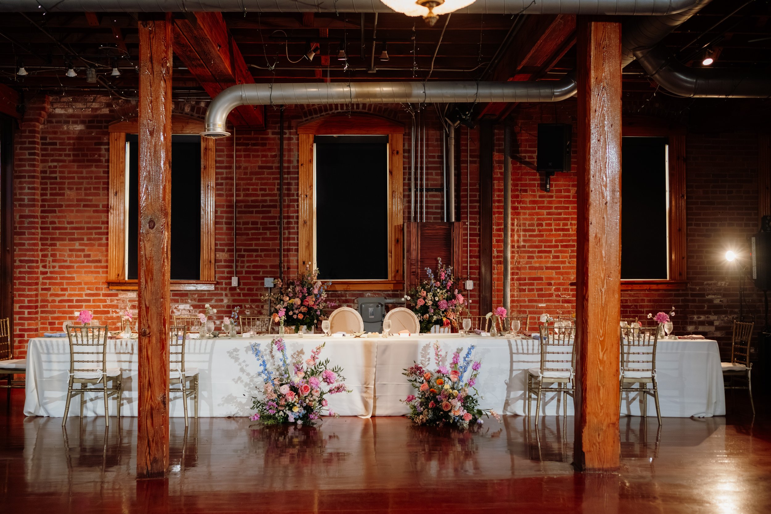 Indoor event space with brick walls, wooden beams, and a large table decorated with colorful flowers, set up for a celebration or wedding reception.
