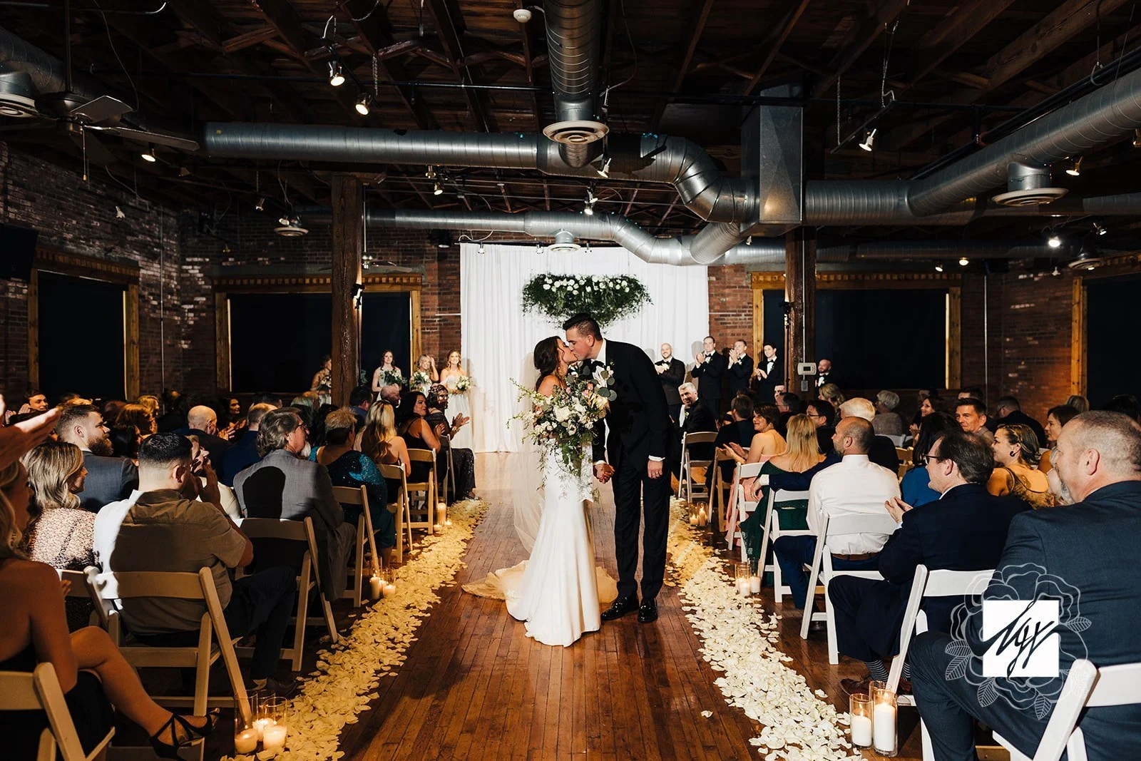 A bride and groom kiss during their wedding ceremony in an industrial-style venue with exposed brick walls and ductwork, decorated with flowers and candles along the aisle, while their guests sit and watch.