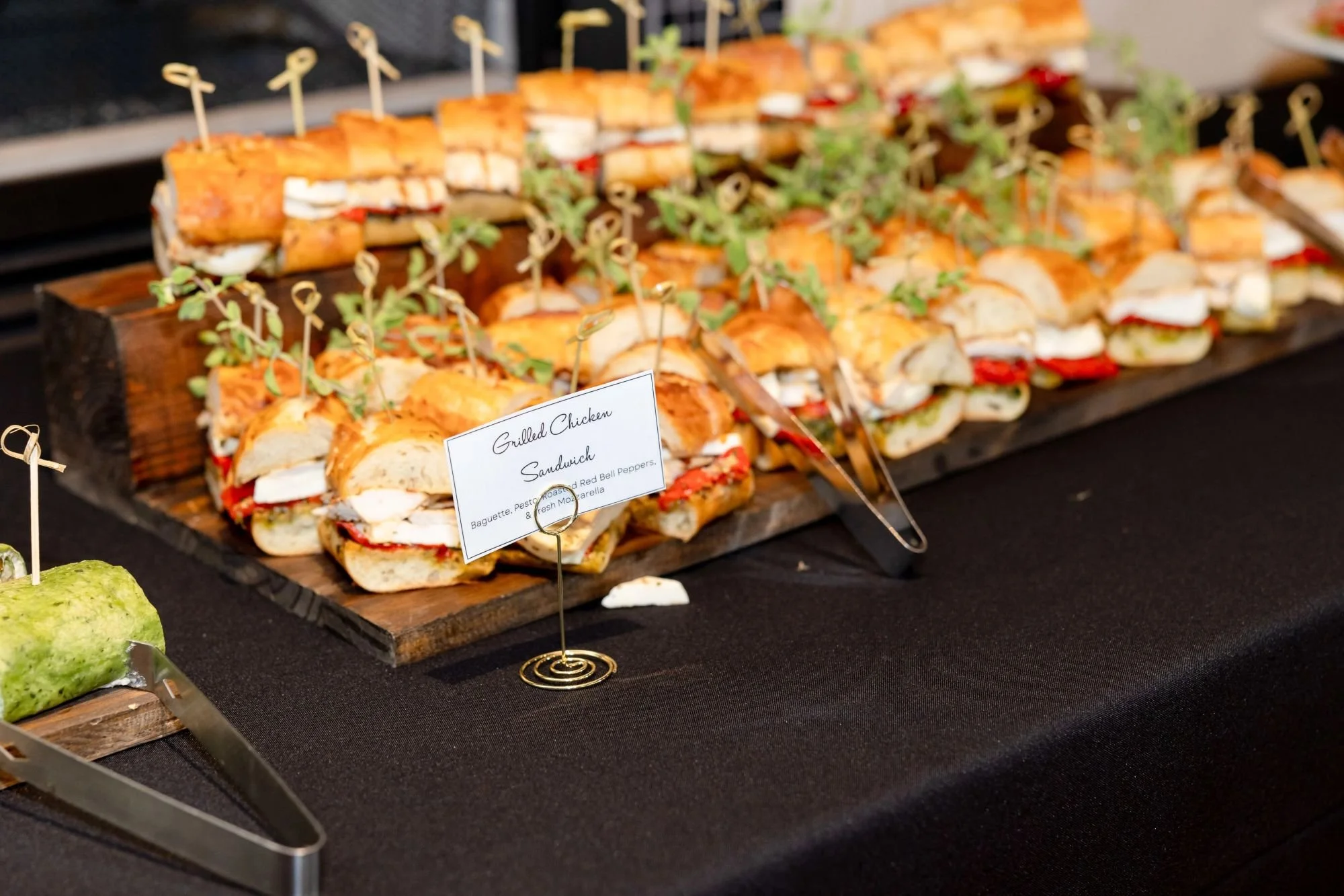 An assortment of small sandwiches, including grilled chicken sandwiches, is displayed on a wooden platter with toothpicks. A small sign indicates they are grilled chicken sandwiches with baguette, roasted red peppers, and fresh mozzarella, garnished 