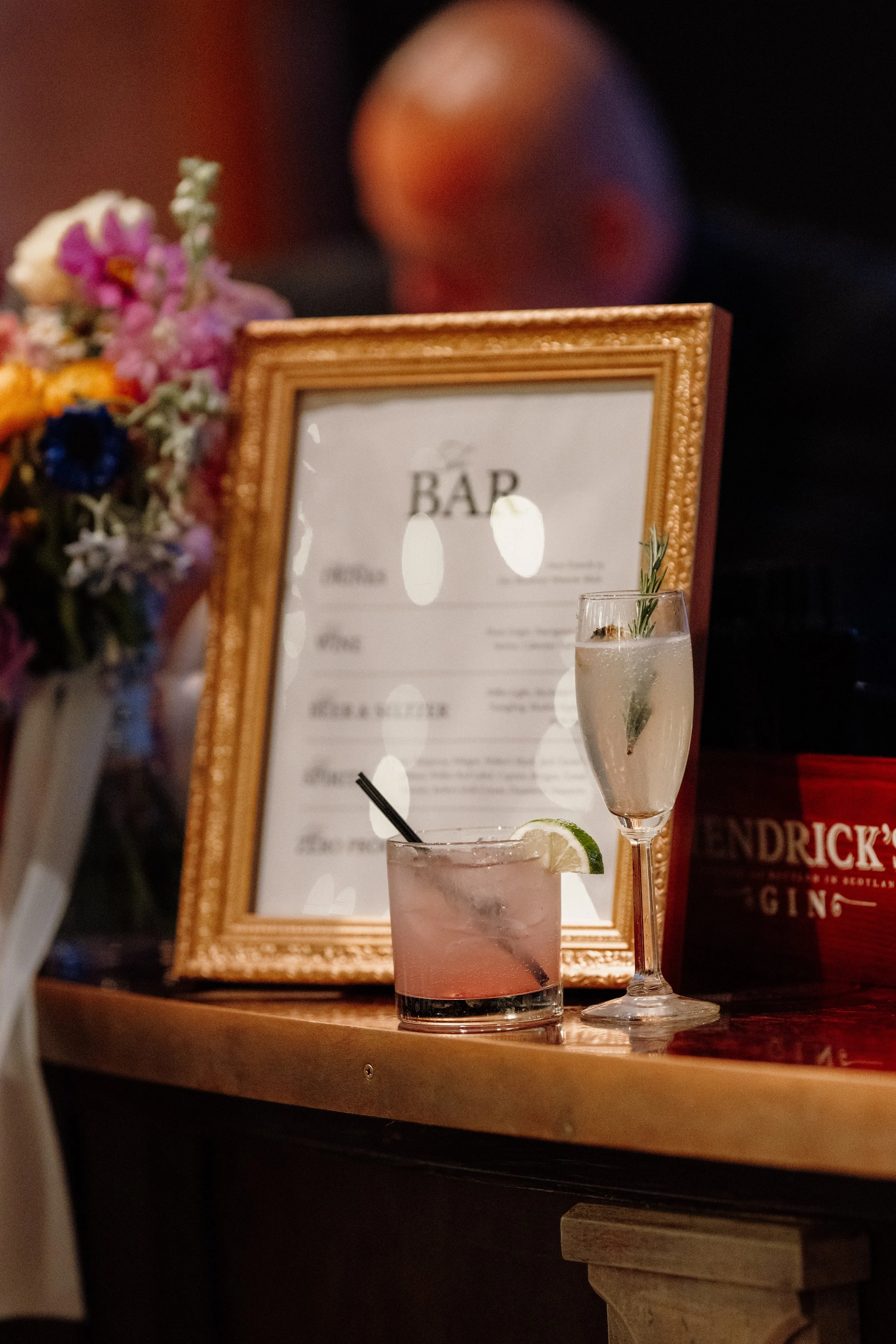 A close-up of two cocktails, one with a slice of lime and a black straw, and the other garnished with a sprig of rosemary, placed on a wooden bar with a decorative gold-framed menu in the background, alongside a blurred floral bouquet.