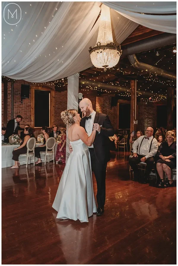 A bride and groom share their first dance at a wedding reception in a decorated venue with draped fabric, fairy lights, and a chandelier.