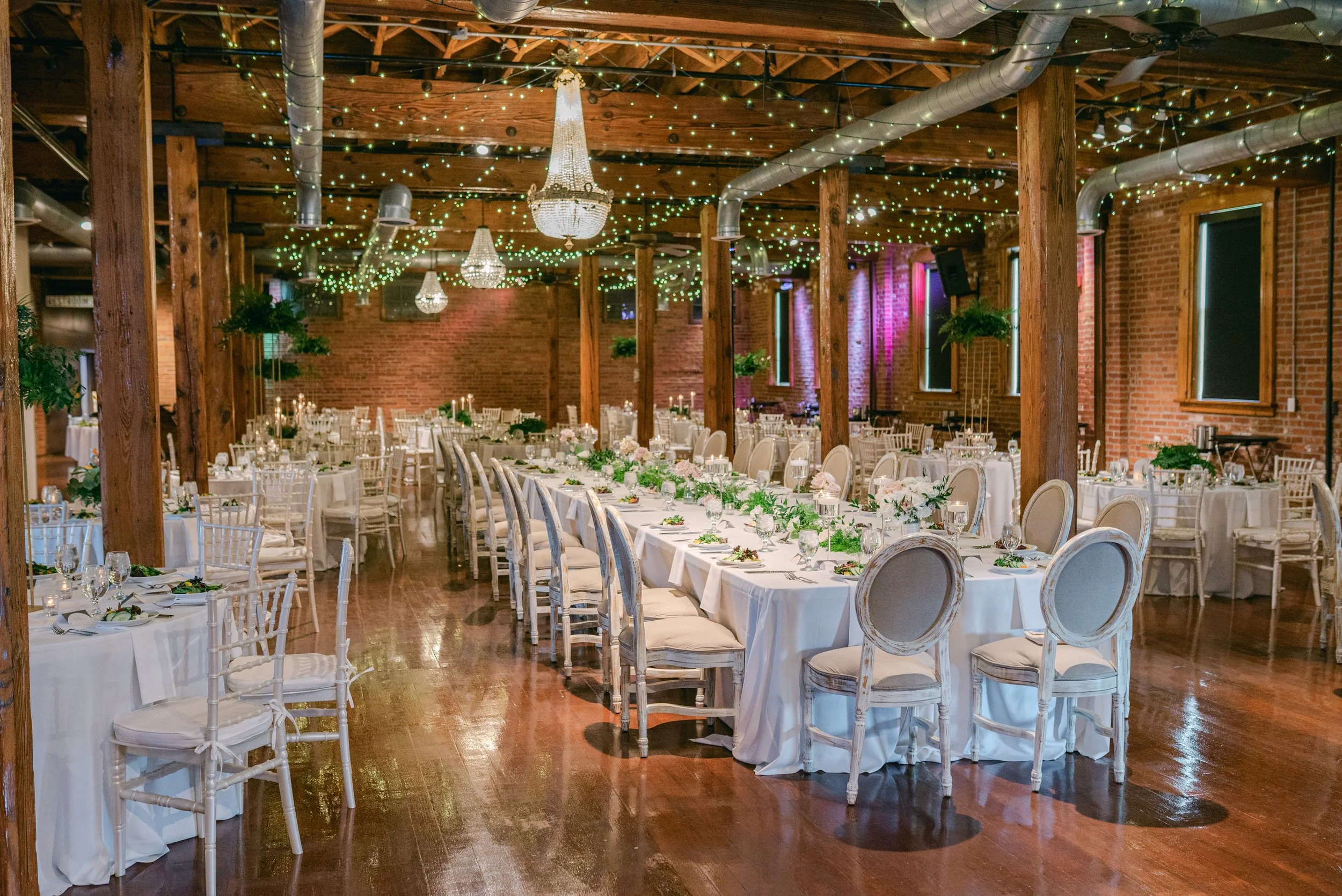 A banquet hall decorated with chandeliers, string lights, and potted plants, with long tables set for a wedding or event, featuring white tablecloths, candles, and floral centerpieces.