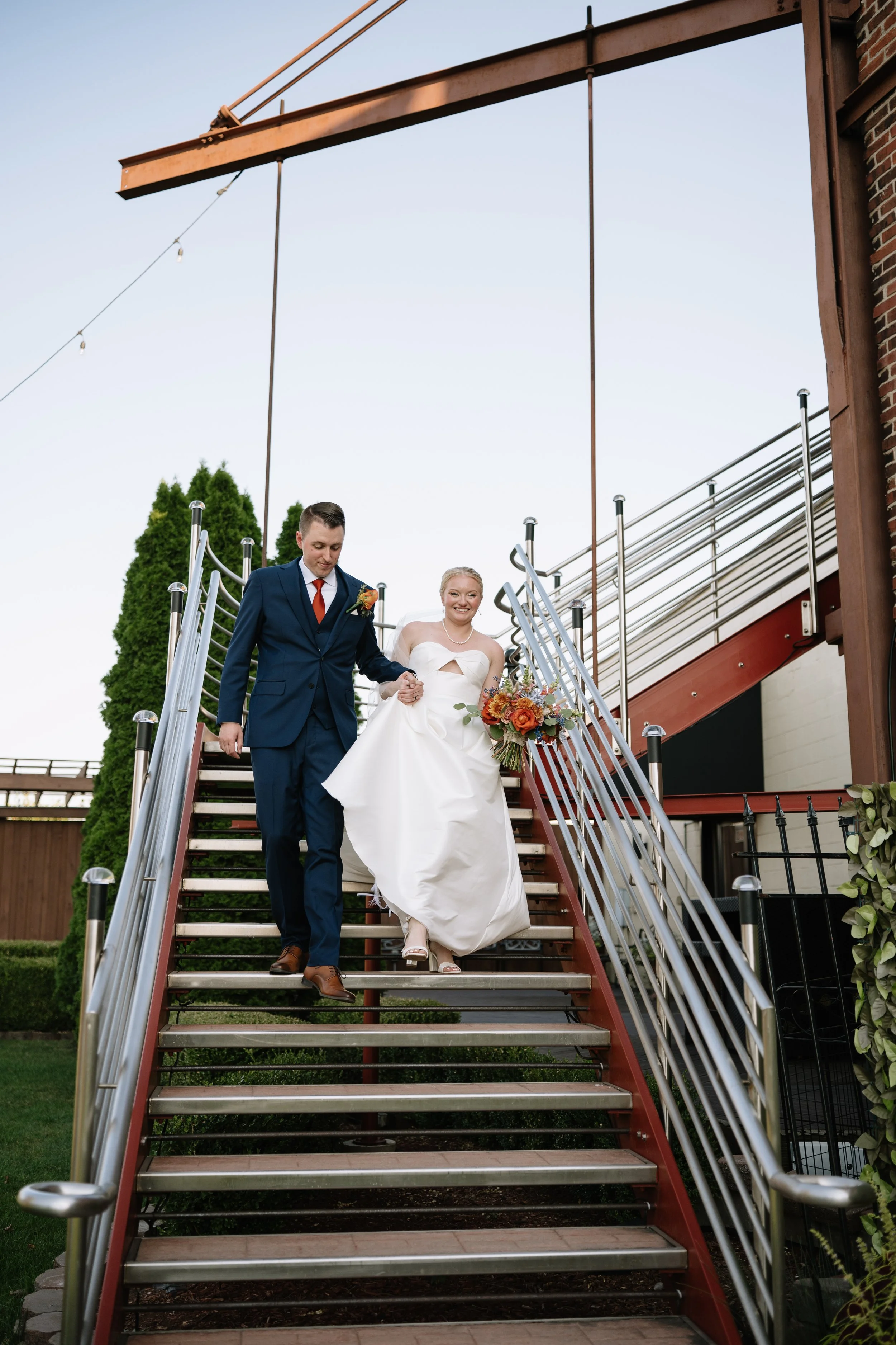 A bride and groom descending outdoor stairs after a wedding ceremony, with the bride holding a bouquet of flowers and smiling.