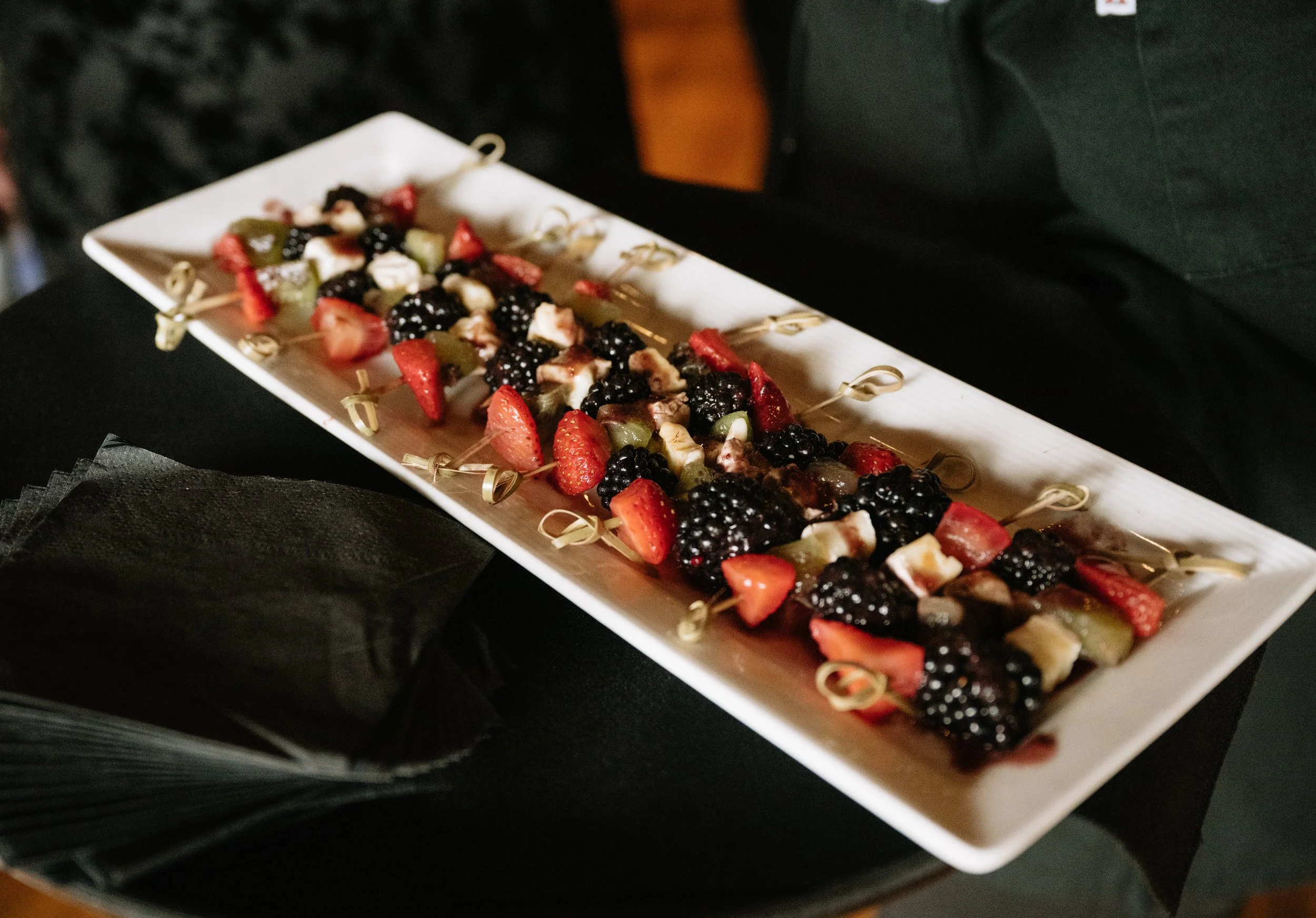 A long rectangular platter with assorted fruit skewers, including strawberries, blackberries, grapes, and possibly cheese cubes.