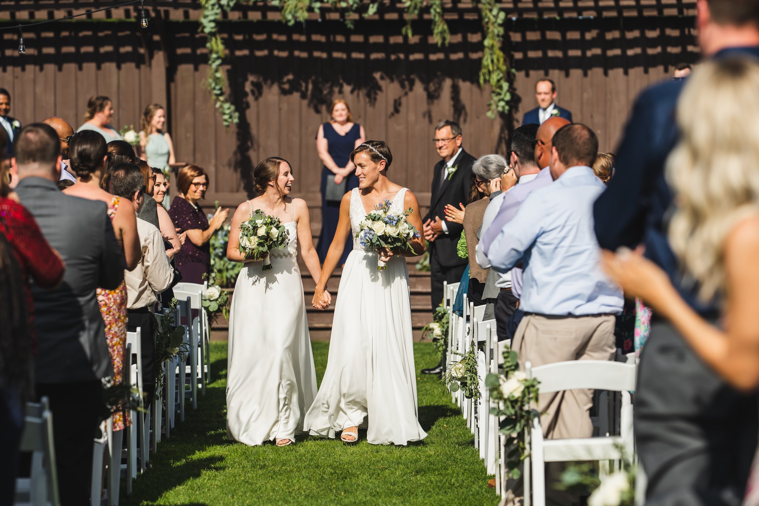 Two women in white wedding dresses are holding hands and walking down the aisle at an outdoor wedding ceremony, smiling at each other. Guests are seated on either side, watching and clapping. The background features a wooden fence and hanging greener