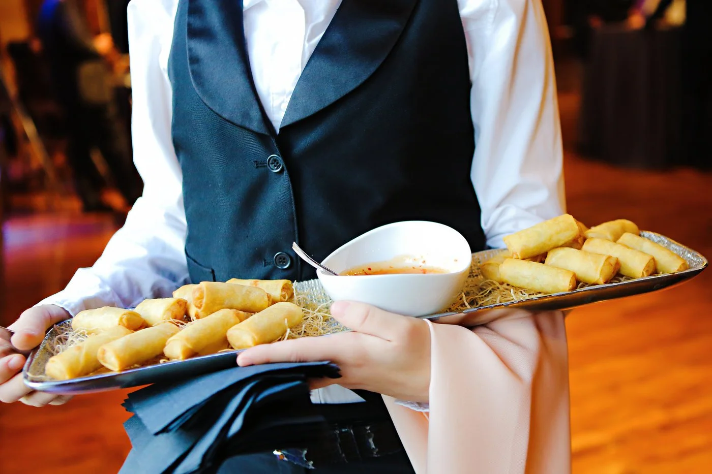 A server in formal attire holding a tray with spring rolls and a bowl of dipping sauce at a restaurant.