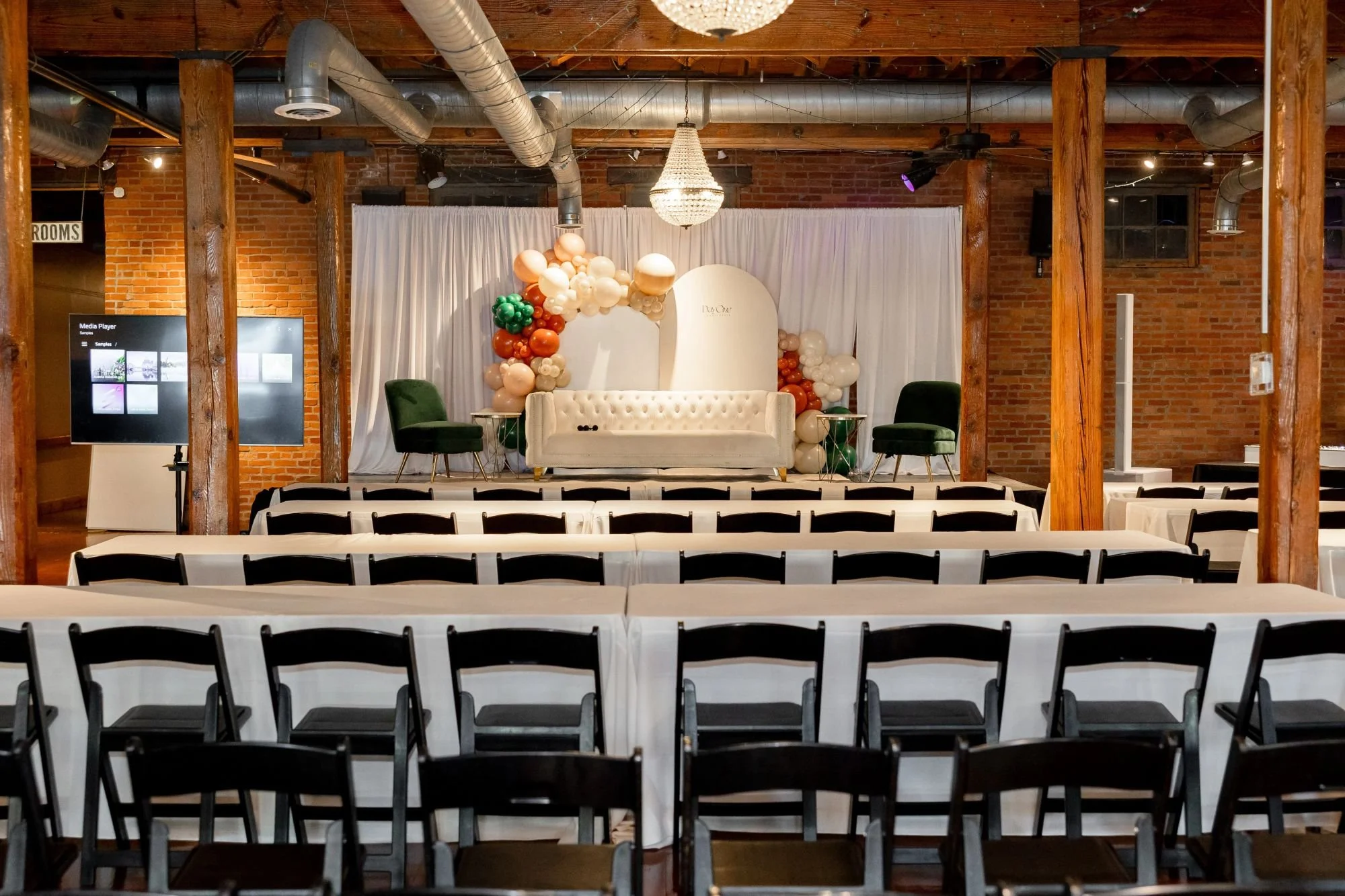 Empty event venue with rows of chairs, a stage with white drapes, balloons, and a chandelier hanging from the wooden ceiling.