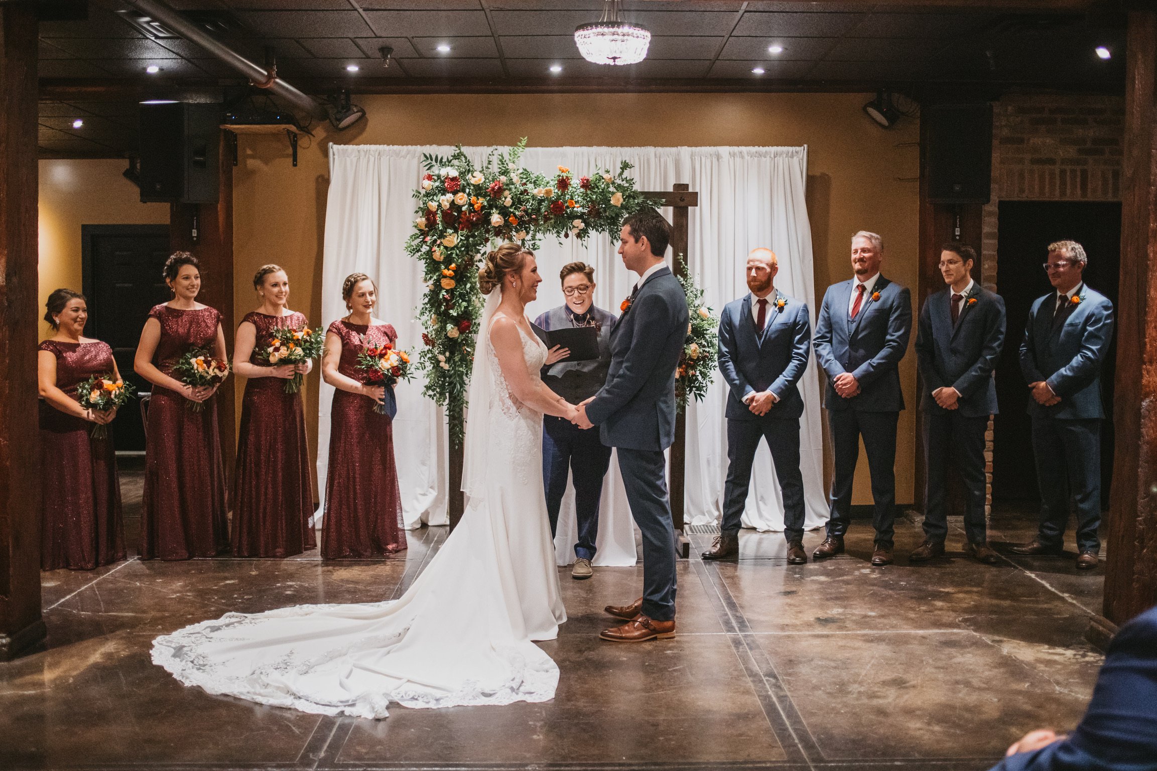 A wedding ceremony in a rustic indoor venue with a bride and groom holding hands and facing each other. The bride wears a white gown with a train, and the groom wears a gray suit. Bridal attendants, including bridesmaids in maroon dresses holding bou