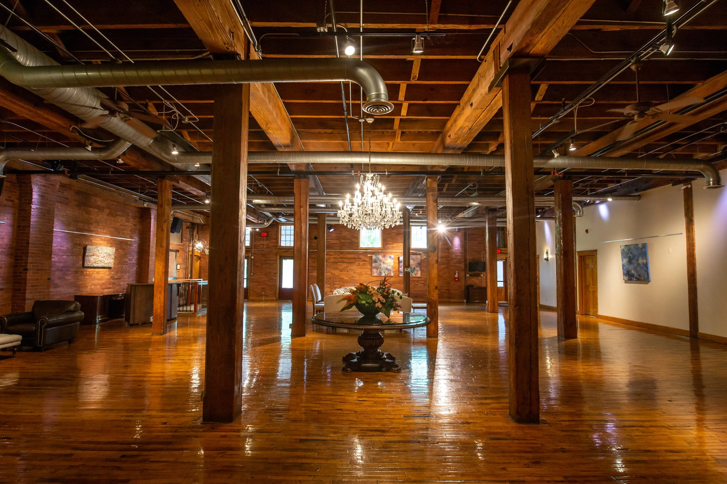 Interior of a spacious, rustic-style event or gathering space with polished wooden floors, exposed brick walls, large wooden beams, and metal ducts on the ceiling. There is a chandelier hanging from the ceiling, and a round table with a floral arrang