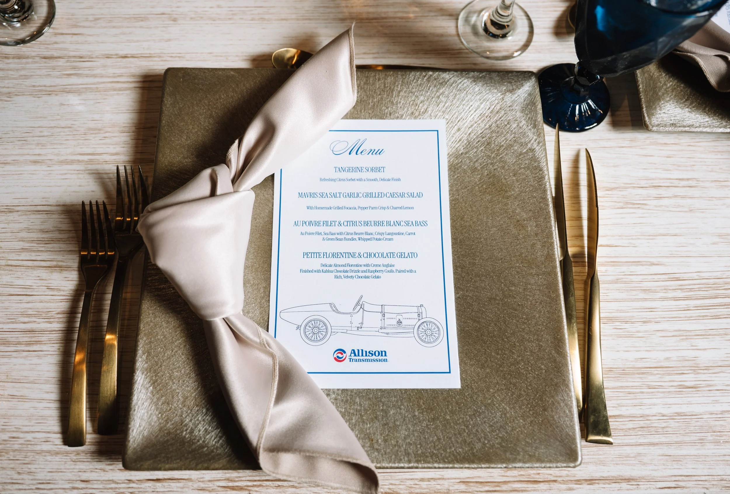 A table setting with a gold charger plate, silver utensils, a white napkin, and a menu featuring seafood dishes, a glass of champagne, and a dark blue wine glass.