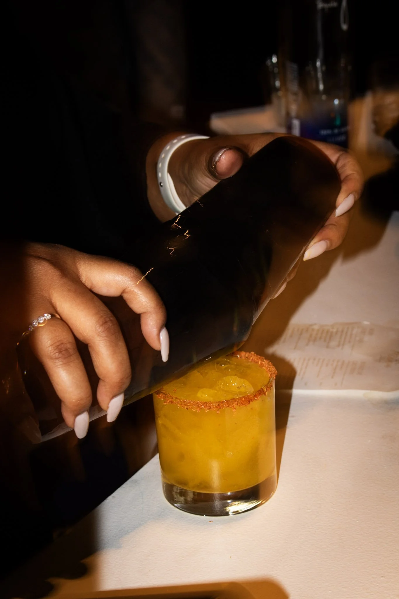 A close-up of a person pouring a yellow drink into a glass with a salted rim and lime wedge, on a white table.