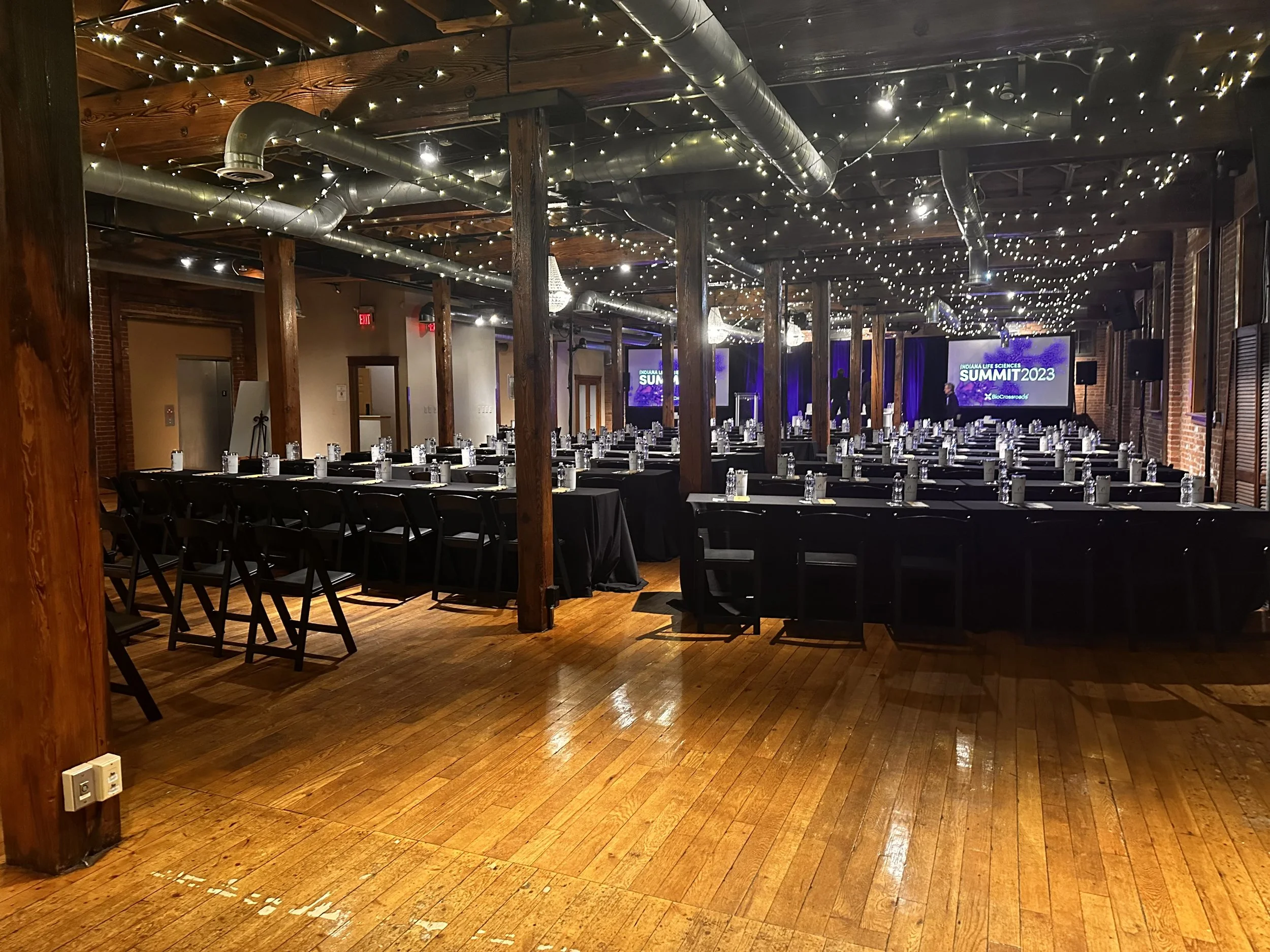 A spacious indoor event venue with wooden floor, decorated with string lights on the ceiling, and set up for a formal event with rows of tables and chairs, water bottles, and a large screen displaying "SUMMIT 2023" in the background.