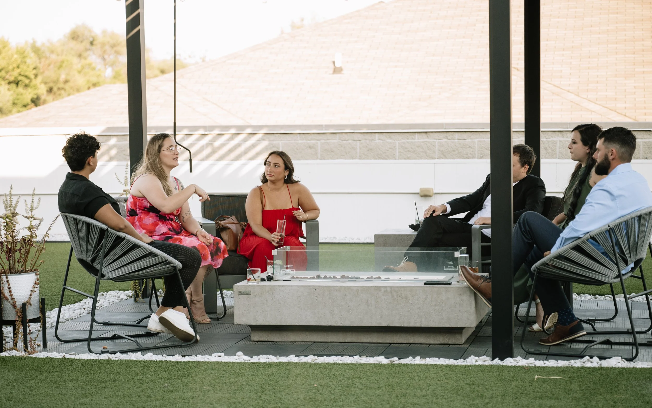 Group of six people sitting and talking on a rooftop patio with modern outdoor furniture and a firepit.
