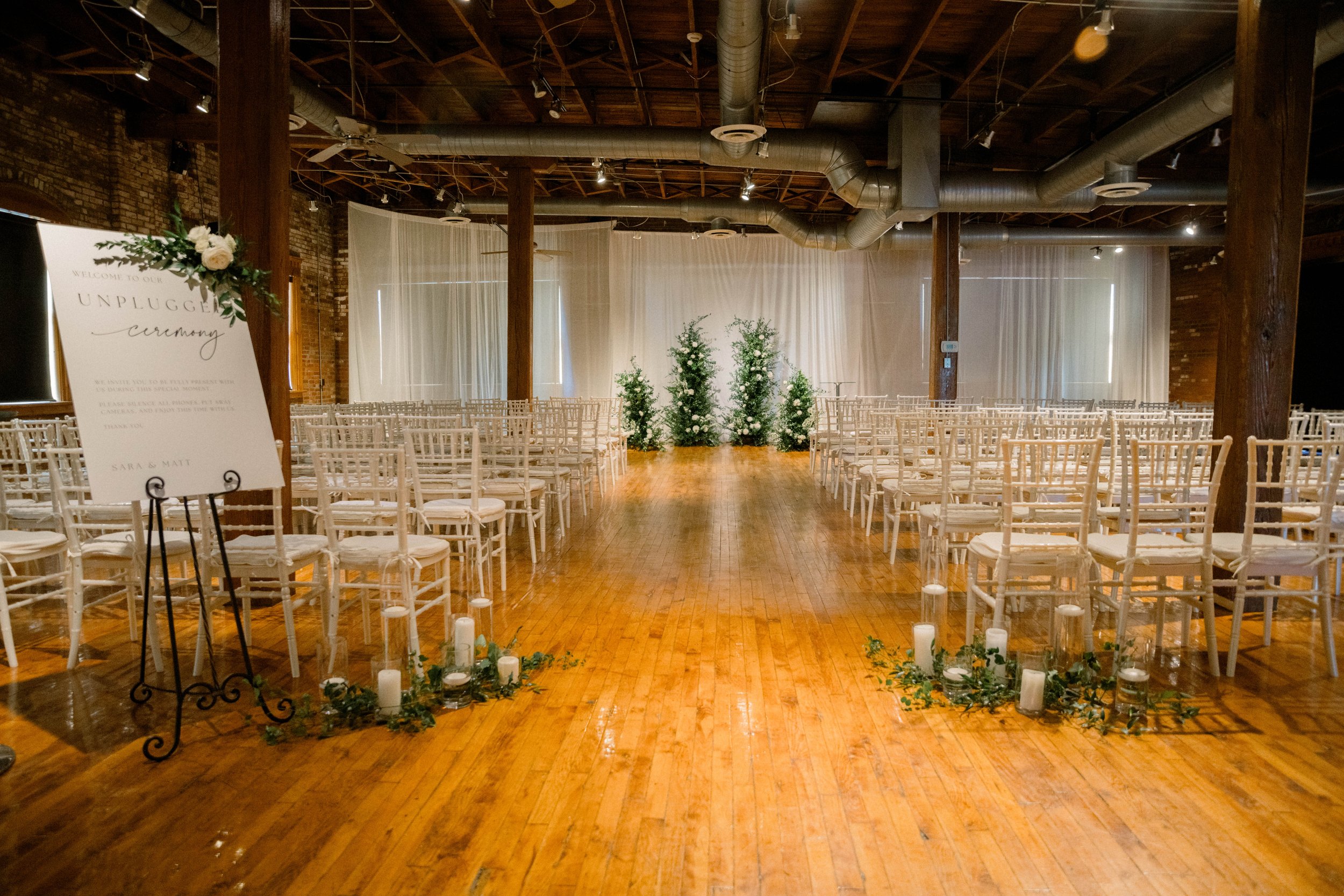 Indoor wedding ceremony setup with rows of white chairs facing an altar with greenery and white flowers, candles along the aisle, with sheer white curtains and a rustic wooden ceiling.