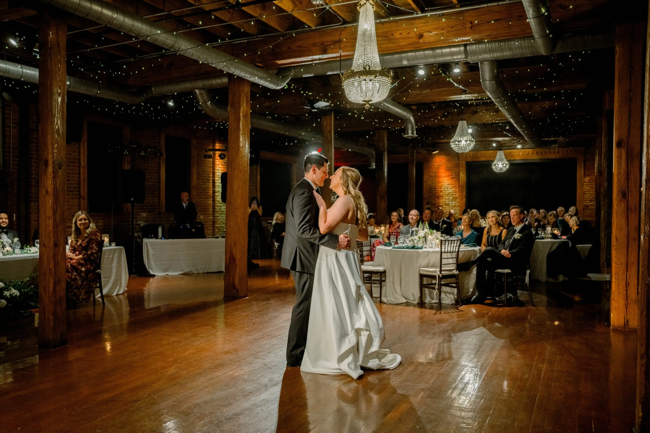 A bride and groom dancing at their wedding reception in a rustic venue with wooden beams and hanging chandeliers, surrounded by seated guests.