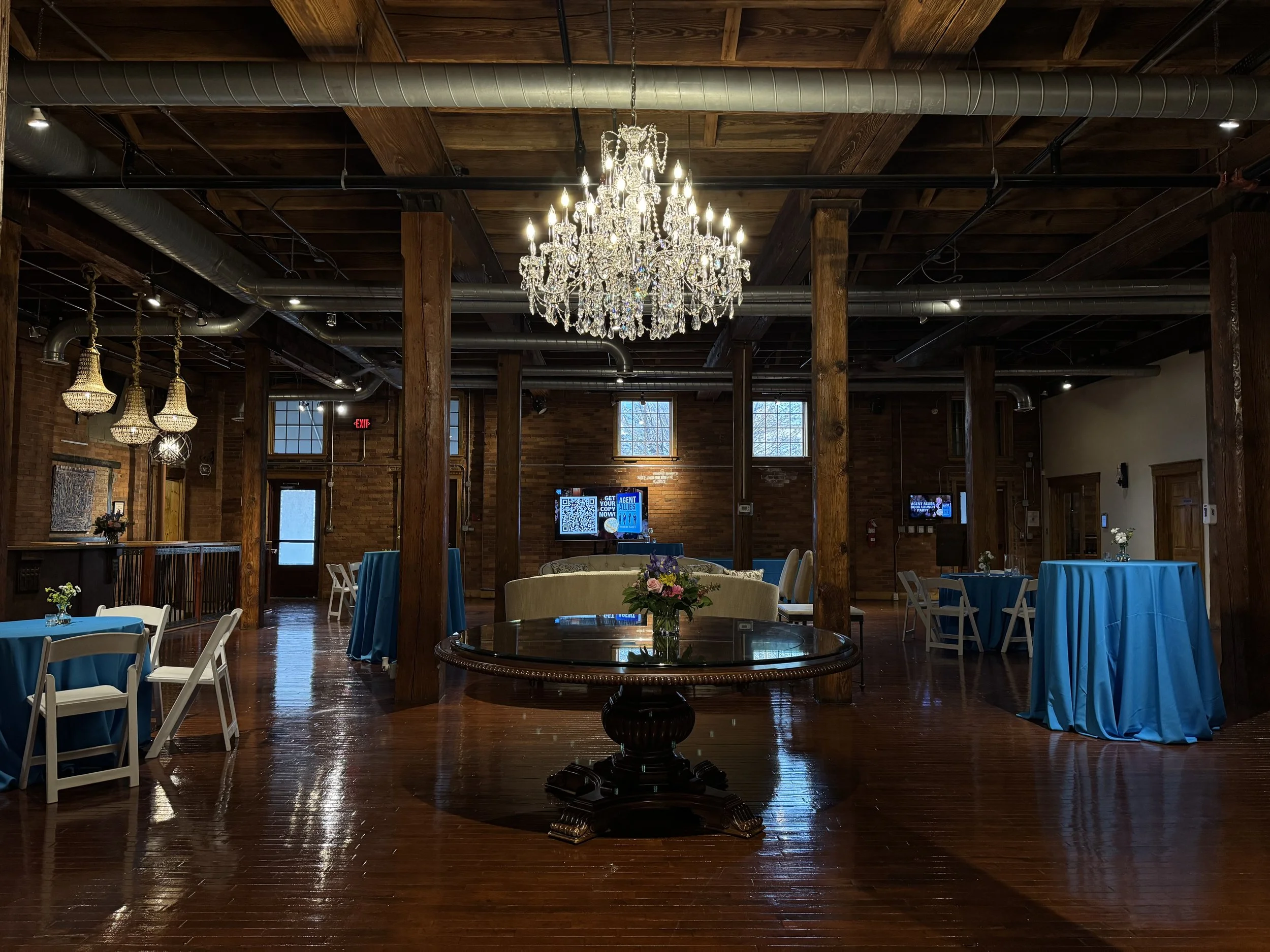 Elegant event space with a large crystal chandelier hanging from a wooden ceiling, surrounded by exposed ductwork. The room has brick walls, wooden columns, and round tables covered with blue tablecloths, decorated with floral centerpieces, and white