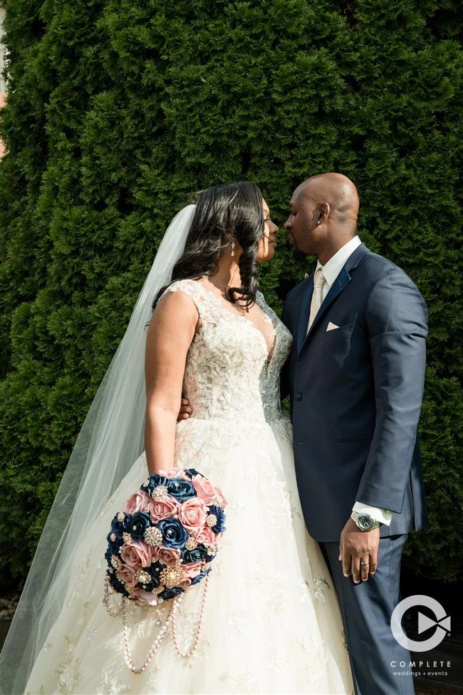 A bride and groom stand close, touching noses, outdoors with a dense green hedge in the background. The bride wears a white lace wedding gown and veil, holding a bouquet of pink and blue roses decorated with pearls. The groom wears a navy blue suit a