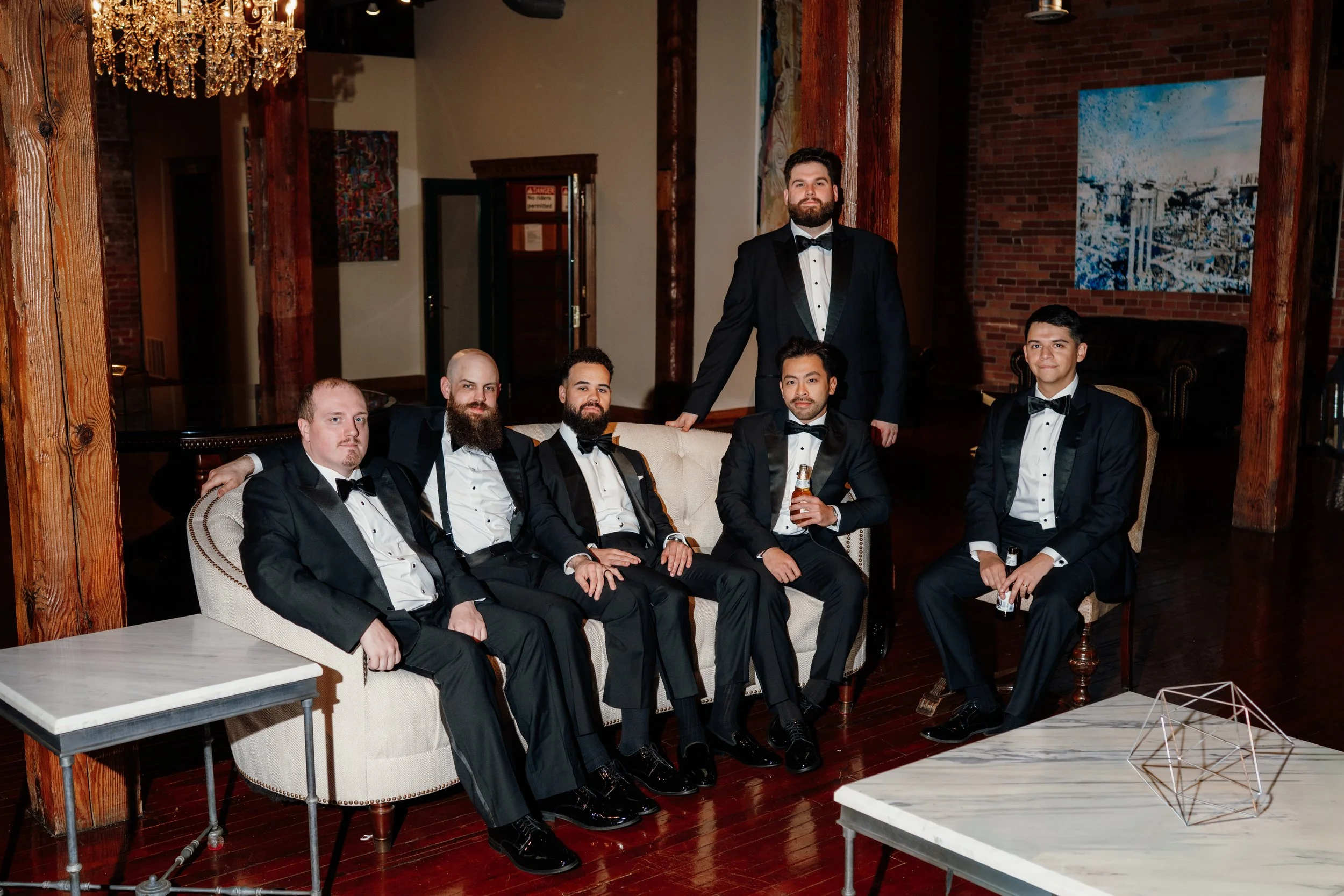 A group of seven men dressed in formal tuxedos sitting and standing on a vintage sofa in a stylish interior with brick walls, wooden beams, and abstract artwork. One man standing behind the sofa holds a drink.