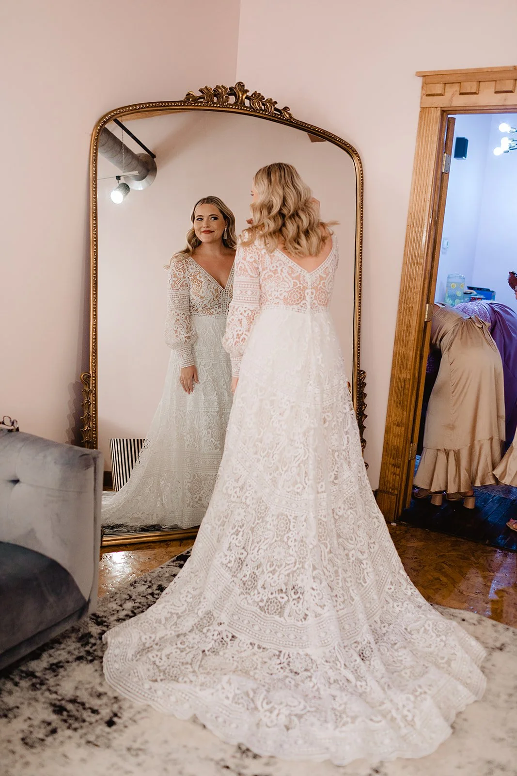 A woman in a white lace wedding dress looking at herself in a large ornate mirror, showing her reflection with soft makeup and blonde hair styled in loose curls.