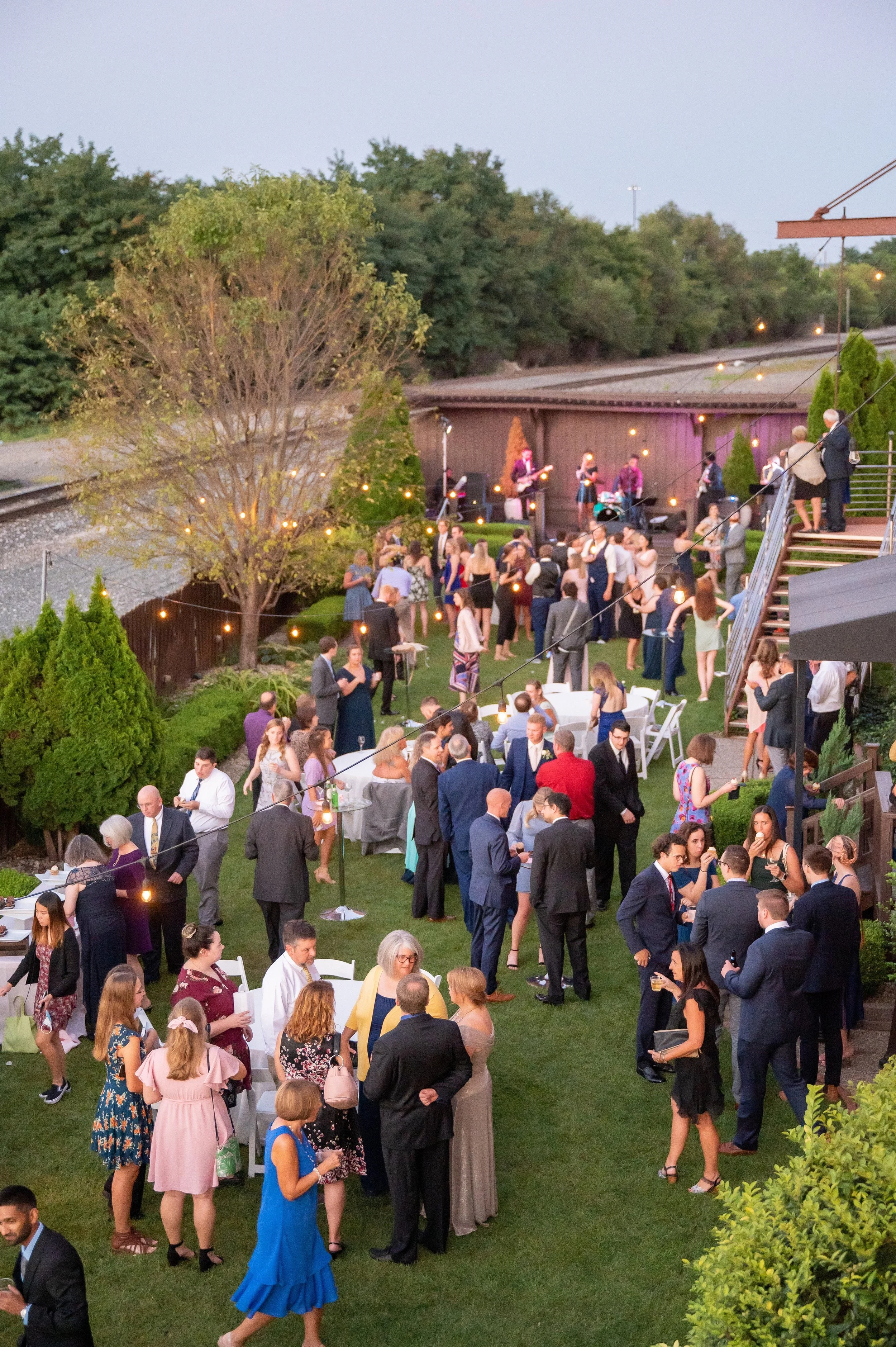 Outdoor event with people socializing, a live band performing on stage, and string lights hanging, set near train tracks and trees as dusk approaches.
