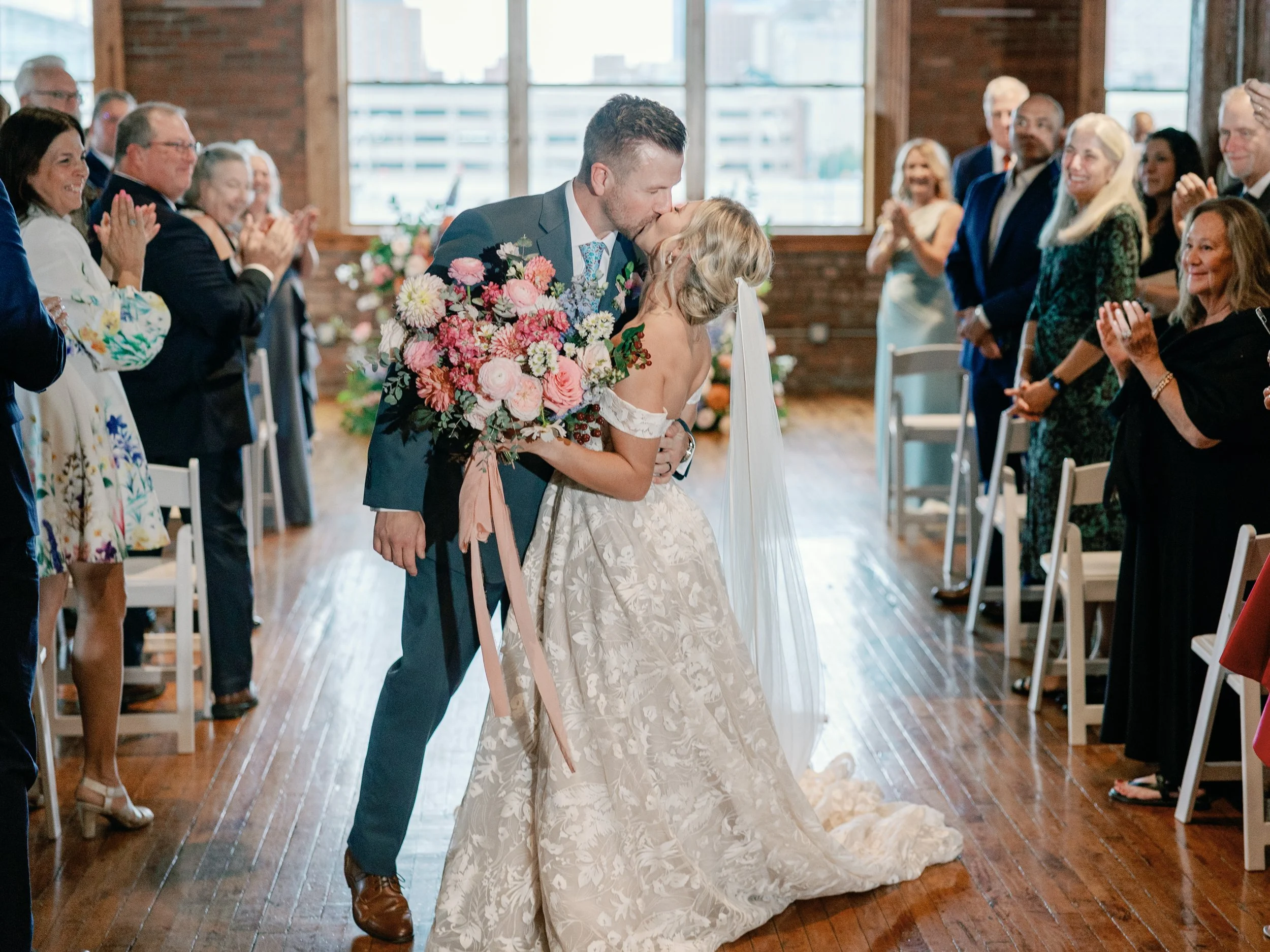 A couple shares a kiss during their wedding ceremony in a wood-floored venue with large windows, surrounded by seated and standing guests clapping and smiling.