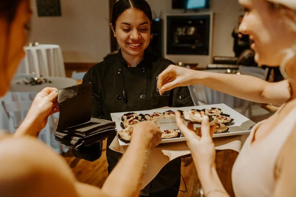 A chef serving a tray of assorted pastries to two women in a restaurant setting.