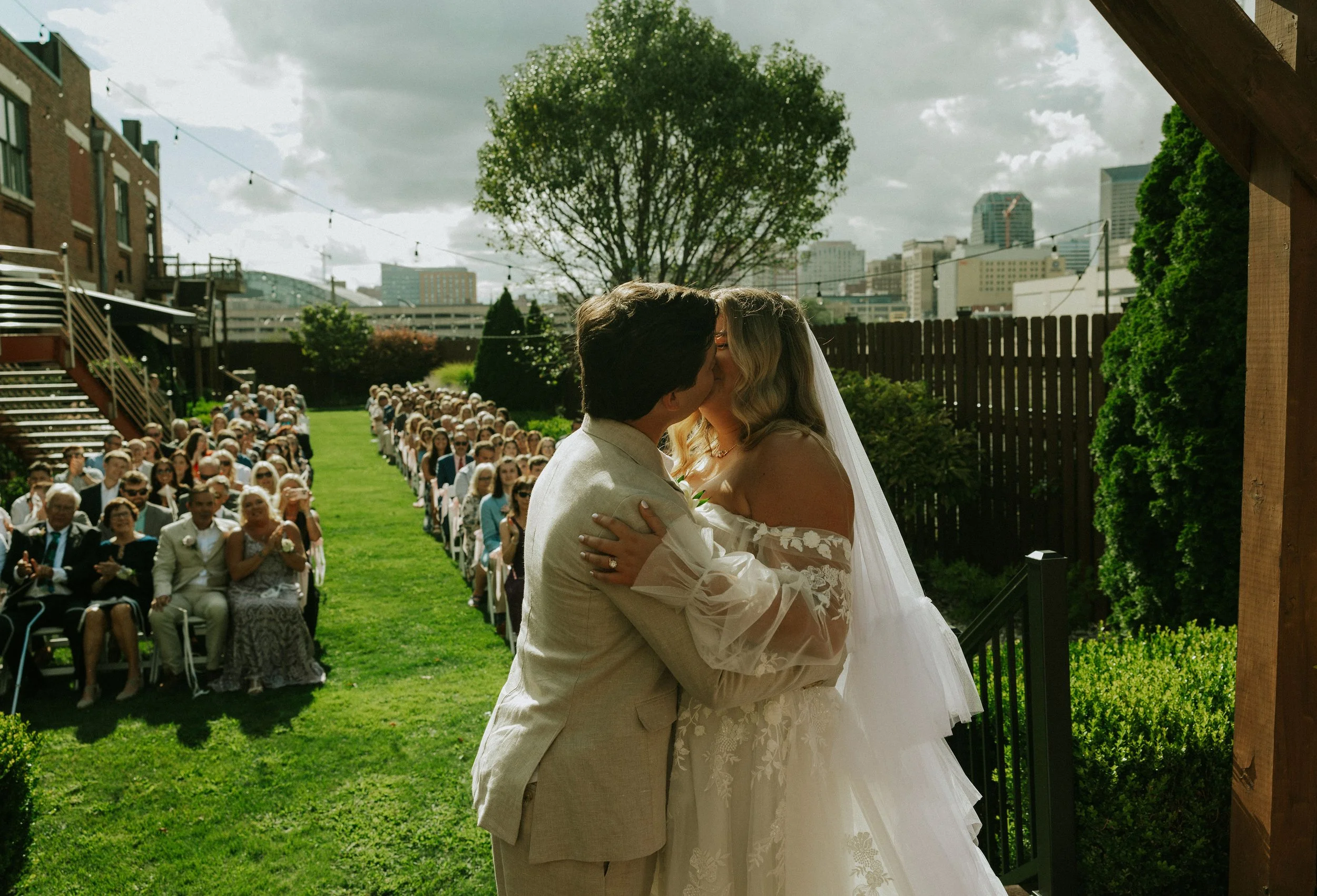 A bride and groom kiss during their outdoor wedding ceremony, with rows of seated guests and a city skyline in the background.