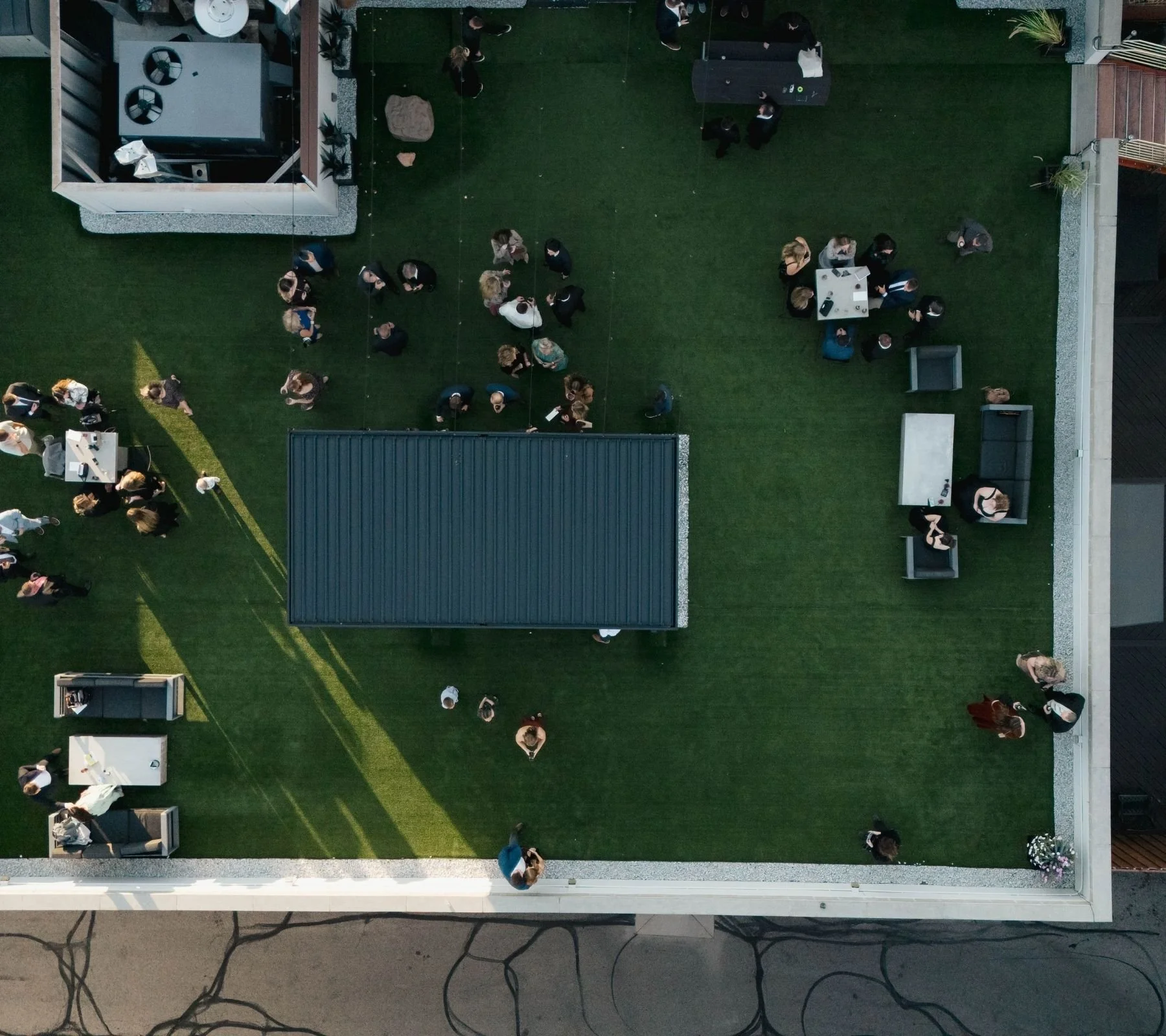 An aerial view of a rooftop garden with a green turf, outdoor furniture, and numerous people mingling at a social gathering.