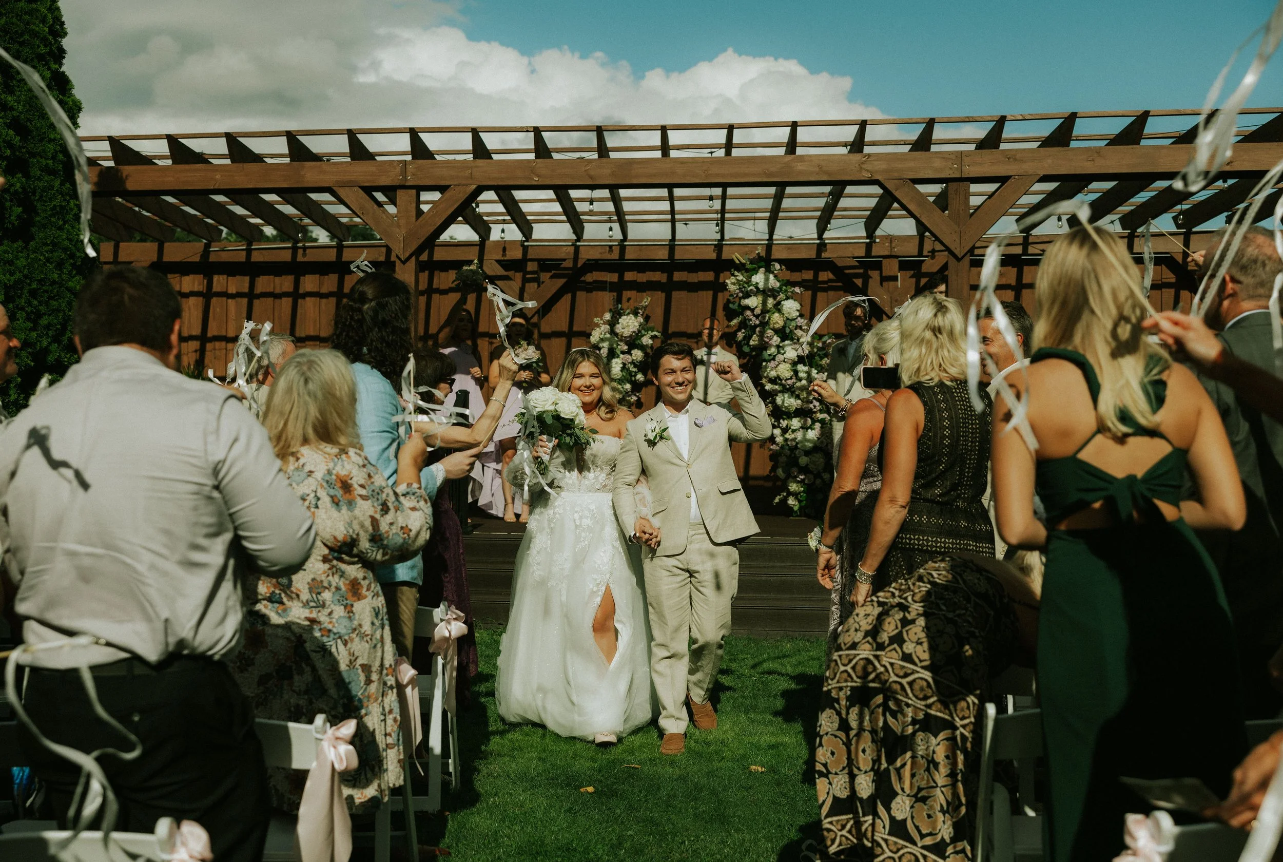A bride and groom walking down the aisle after their wedding ceremony, surrounded by guests celebrating outdoors under a wooden structure on a sunny day.