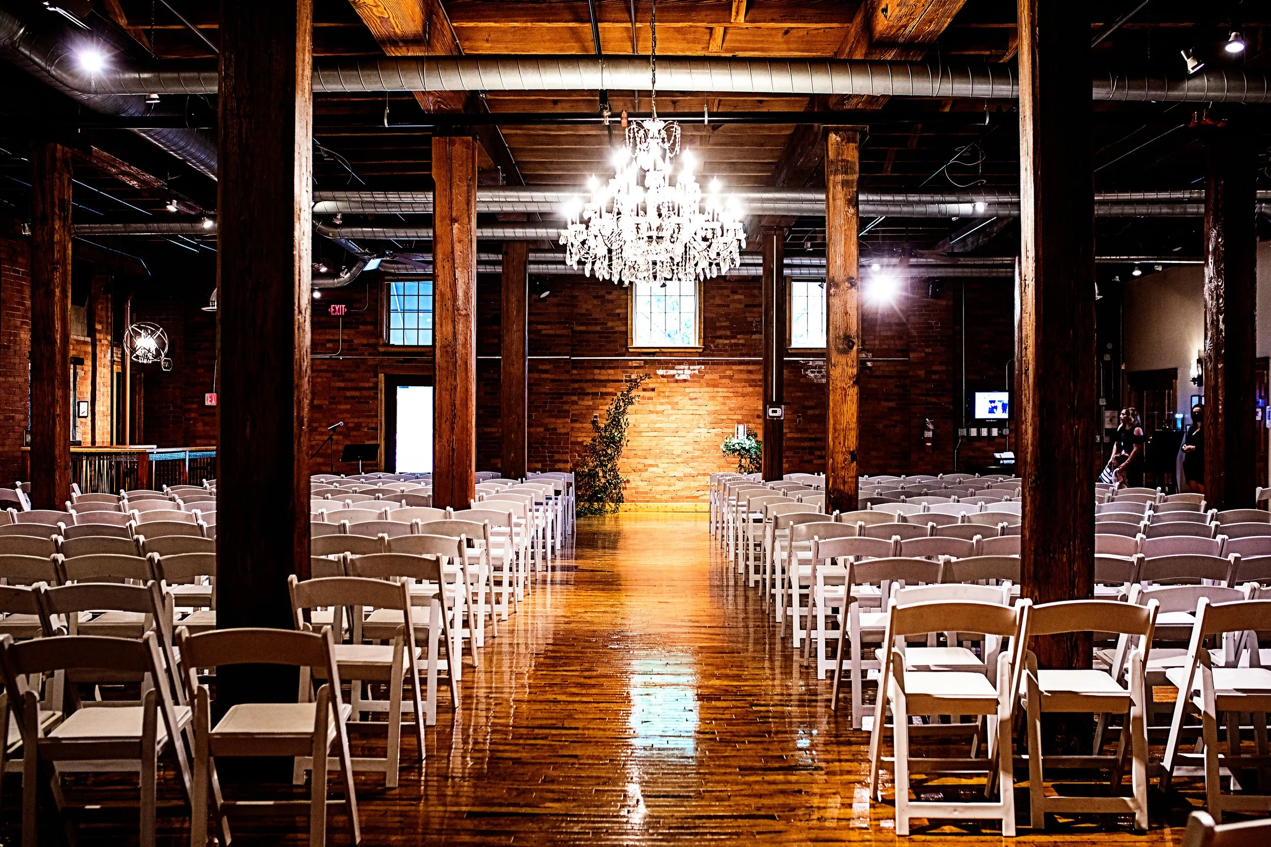 Interior of a spacious venue or hall with rows of white chairs, a large chandelier hanging from the wooden ceiling, brick walls, and natural light coming through windows.