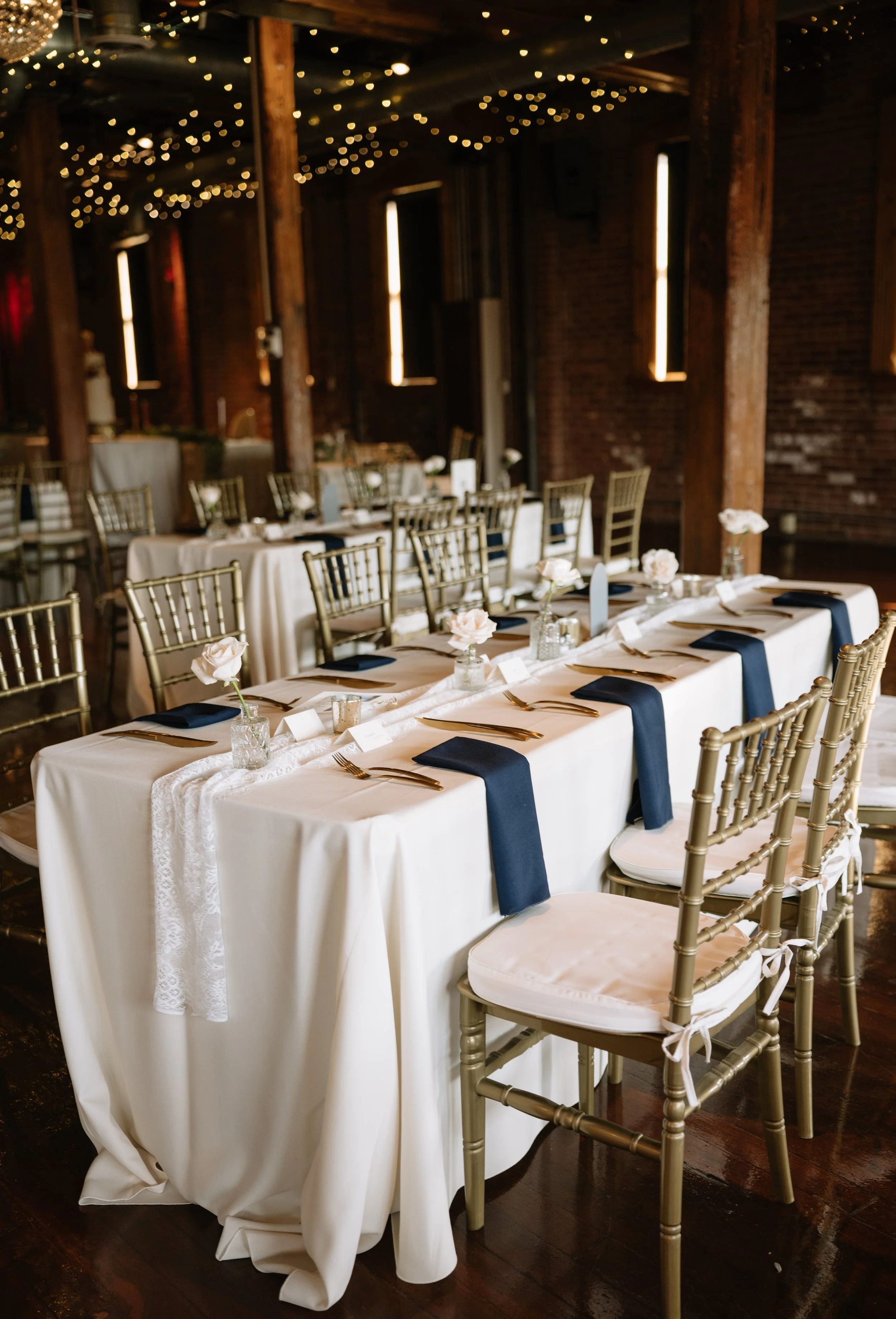 Wedding reception setup with long table covered with white tablecloth, decorated with small vases of white flowers, navy blue napkins, and gold utensils, in a rustic style venue with string lights hanging from wooden beams.